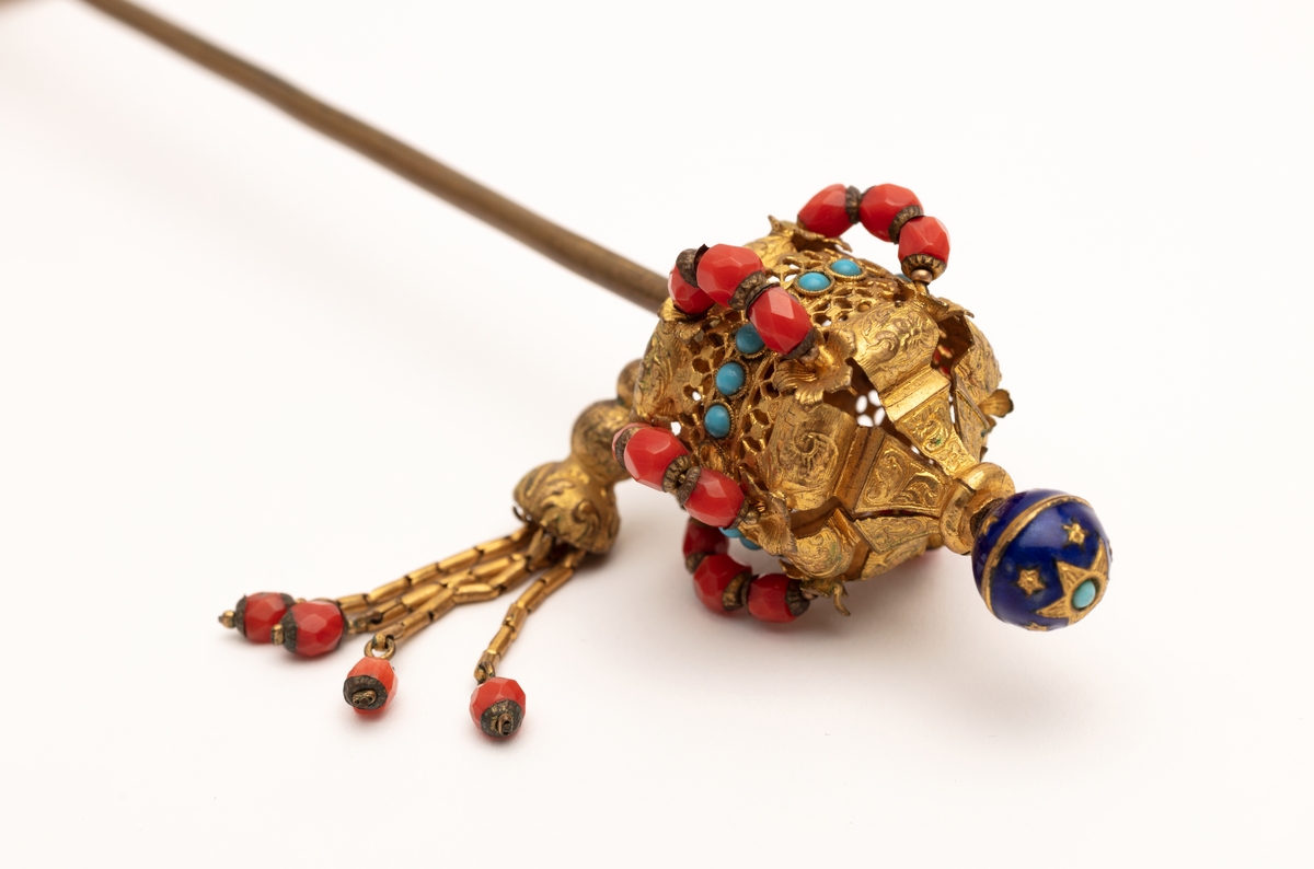 Hattenål i metall, delvis forgylt. Endestykkene er dekorert med røde og blå glassperler og dusker.