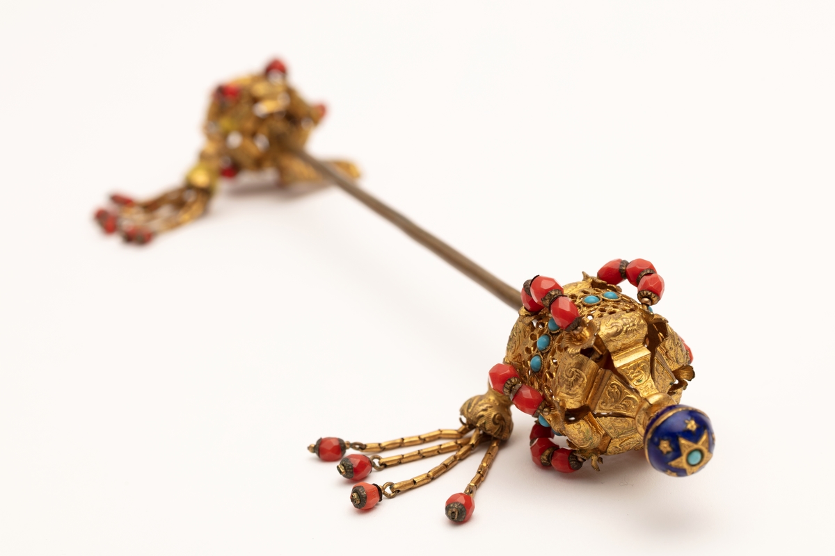 Hattenål i metall, delvis forgylt. Endestykkene er dekorert med røde og blå glassperler og dusker.