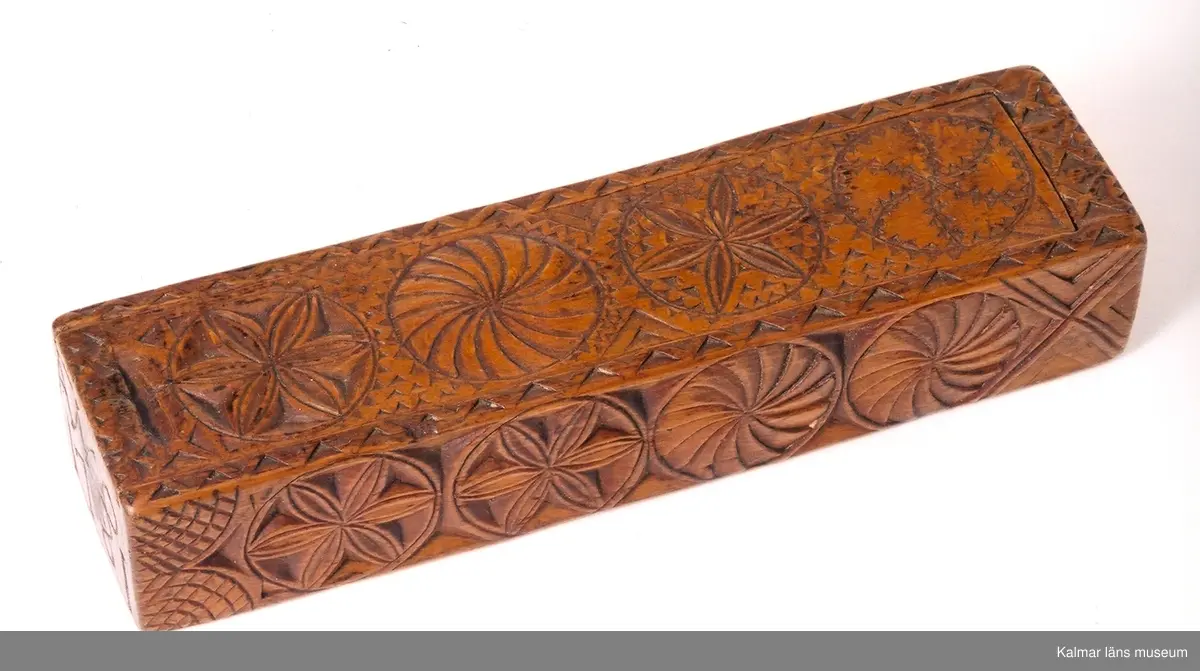 KLM 6626. Raklåda av björk. Raklådan skuren ur ett stycke. Skjutlock av bok. Lock och långsidor ornerade med karvsnittsrosetter och virvelhjul. På ena gaveln inskuret ANO 1781, på den andra ett LN 52, (LN ihopskriver). I bottens undersida inslagna mässingsstift bildande initialerna LN. Lådan fernissad i senare tid.