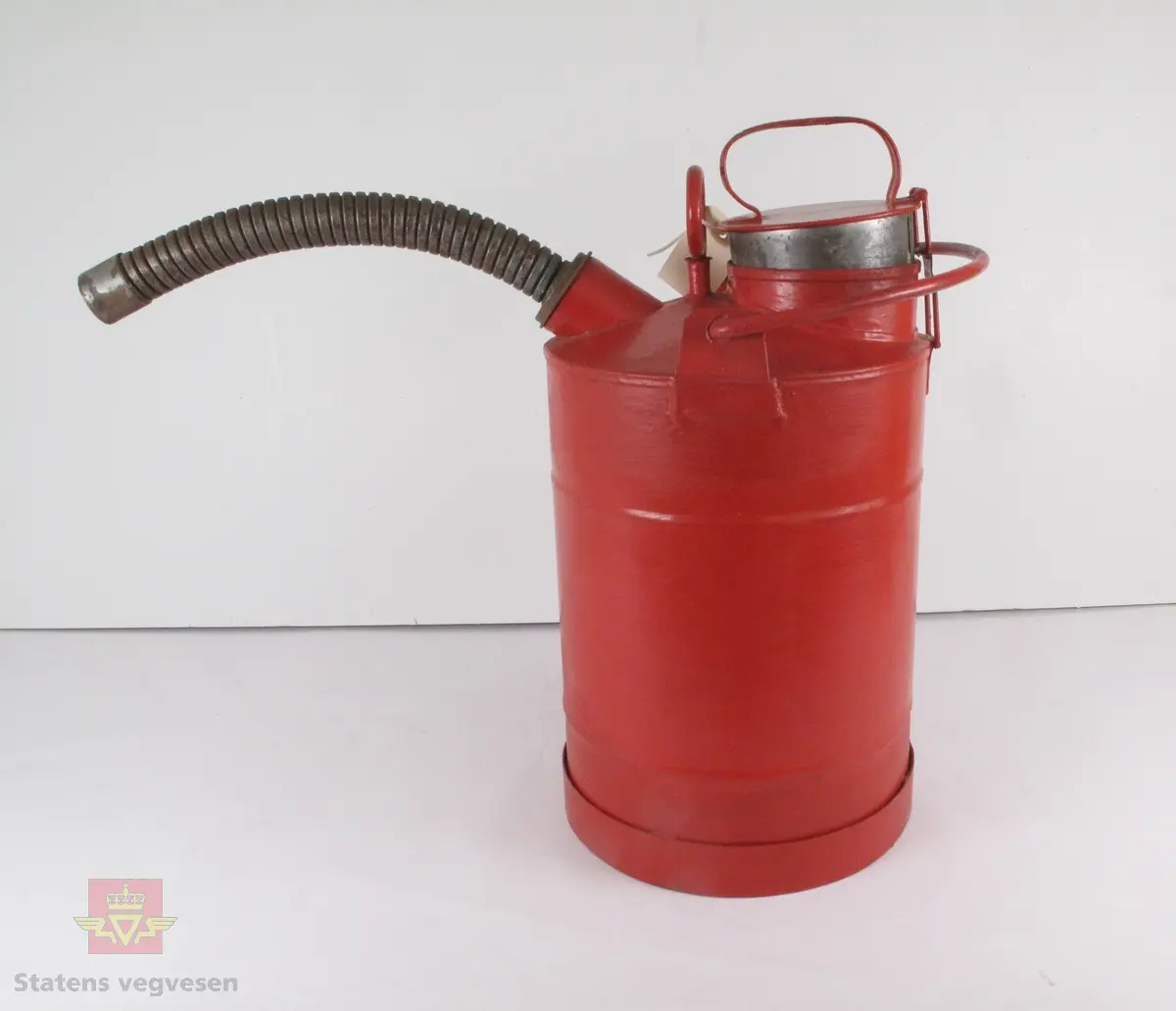 Rød oljekanne med håndtak på lokket der man kan fylle på olje, samt et håndtak som er formet som en bøyle til hele kannen. En tut i metall sitter i helleåpningen.