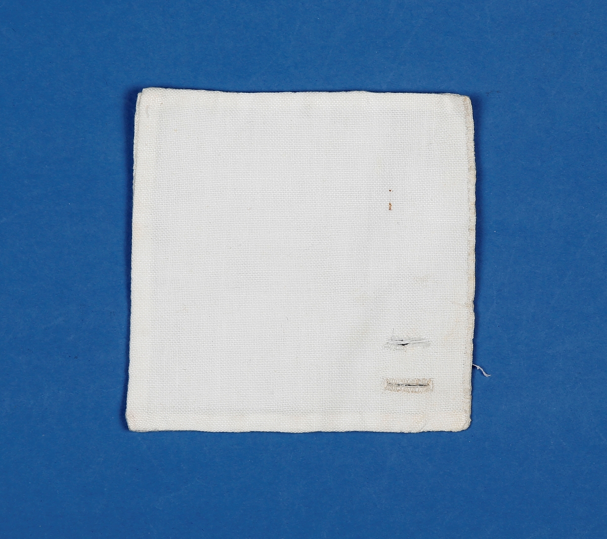 Kvadratisk lapp i litt grov bomull med et ferdig og et påbegynt knapphull sydd for hånd. Lappen er vrangsydd på tre kanter og sydd igjen med knapphulls sting på den fjerde, fra retten.
