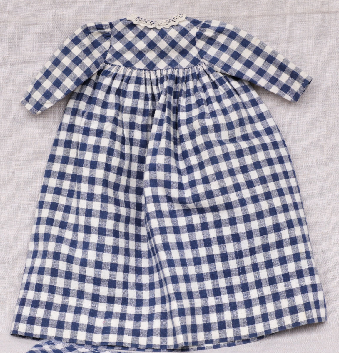 Tre enkle dukkekjoler, to rødprikkete (a og b) og en blårutet (c), en stråhatt (d), et bånd (e). Rød kjole (b) uten ermer, med hvite kanter.