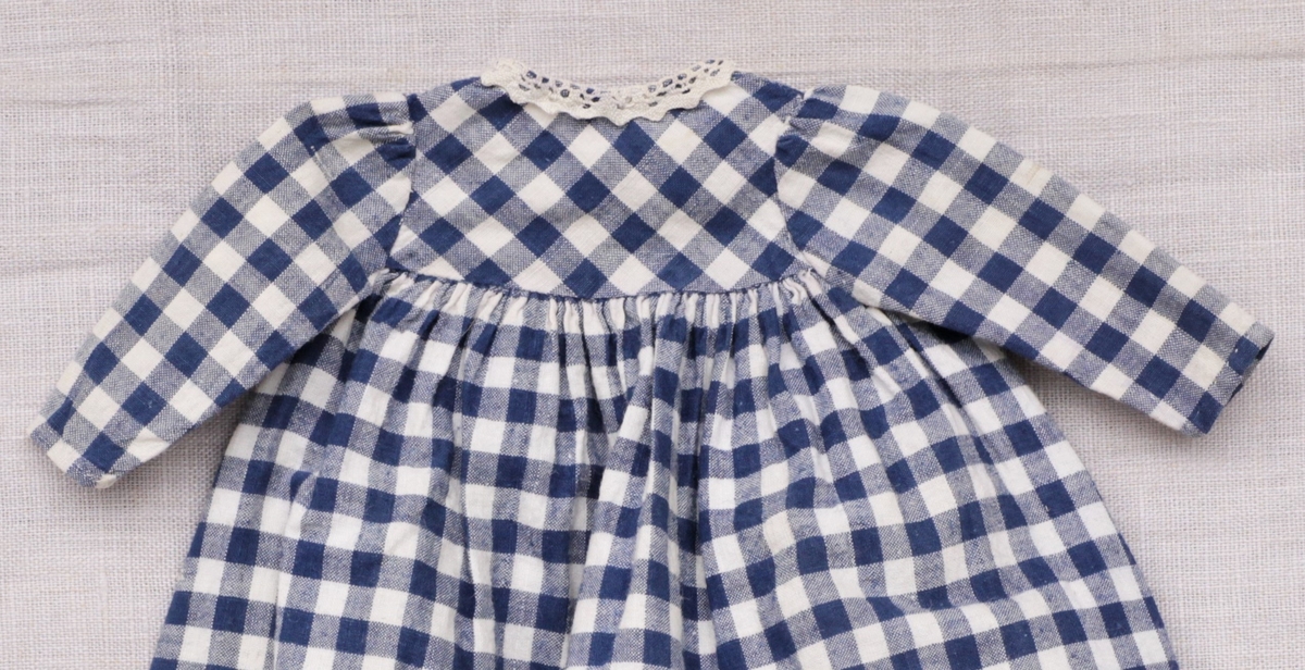 Tre enkle dukkekjoler, to rødprikkete (a og b) og en blårutet (c), en stråhatt (d), et bånd (e). Rød kjole (b) uten ermer, med hvite kanter.