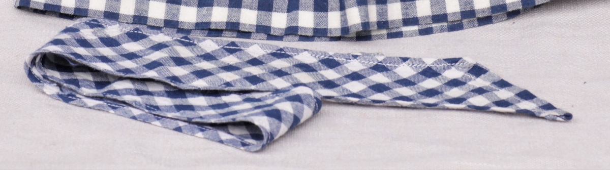 Tre enkle dukkekjoler, to rødprikkete (a og b) og en blårutet (c), en stråhatt (d), et bånd (e). Rød kjole (b) uten ermer, med hvite kanter.