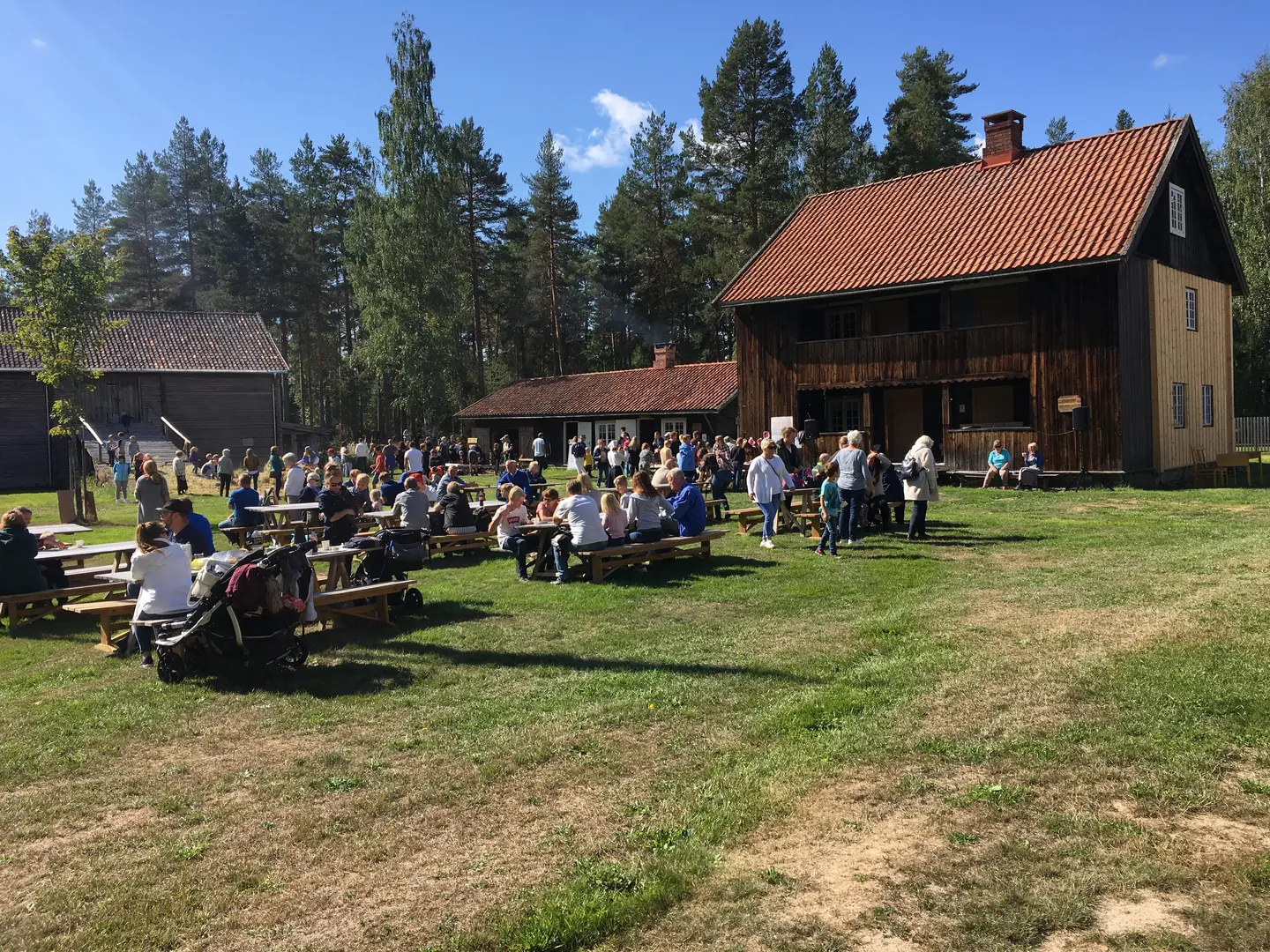 Folk på tunet under Liv i stuene. Fint sommervær. Mange mennesker sitter på benker ved bord plassert midt på tunet. På fotografiet ser vi Skjeppestad, drengestua og låven.