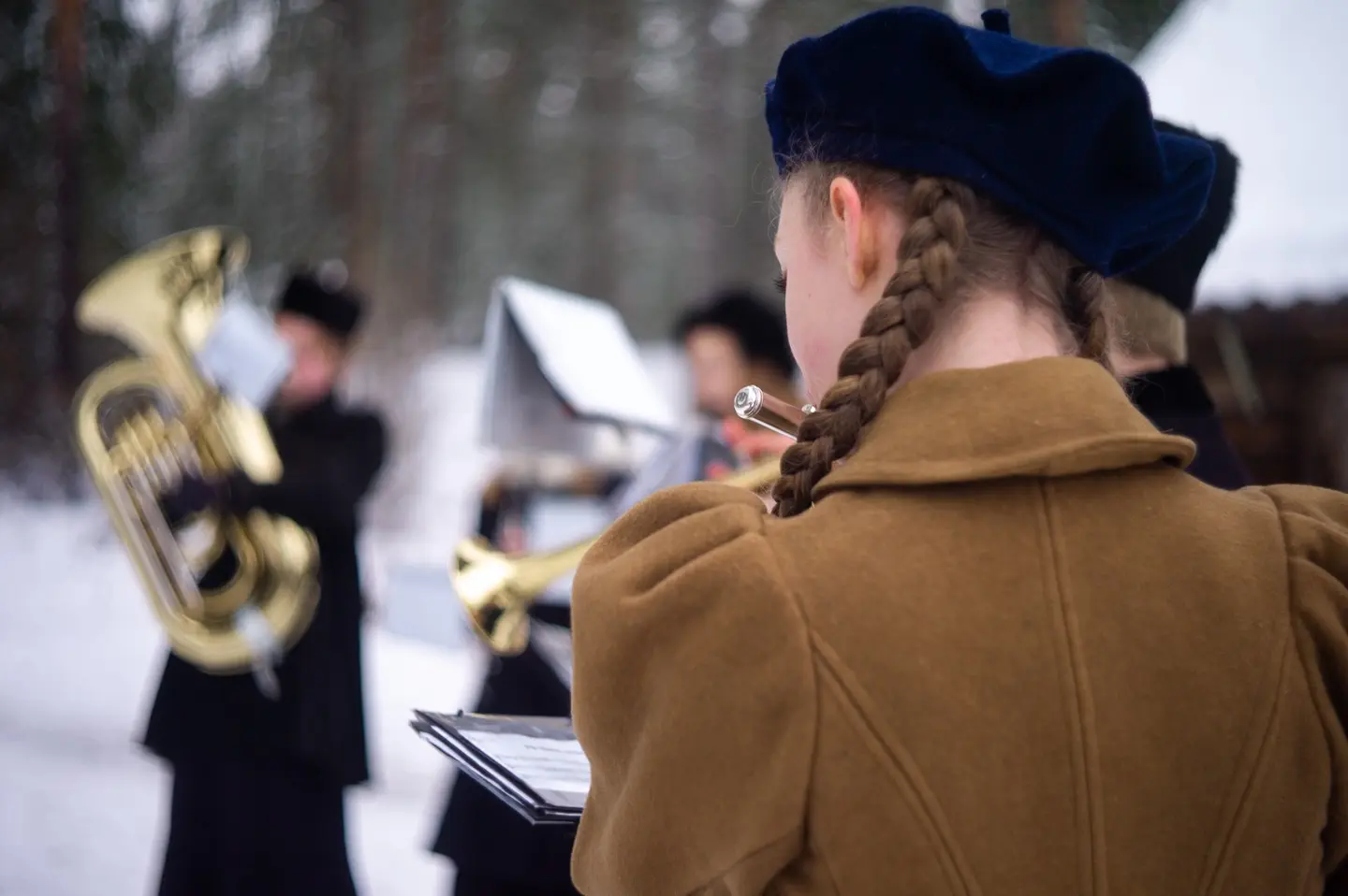 Jul i stuene. Fotografiet viser et skolekorps kledd anno år 1900 som spiller musikk under Jul i stuene. En jente med lysebrun kåpe, blå lue og med musefletter som spiller fløyte er i fokus.