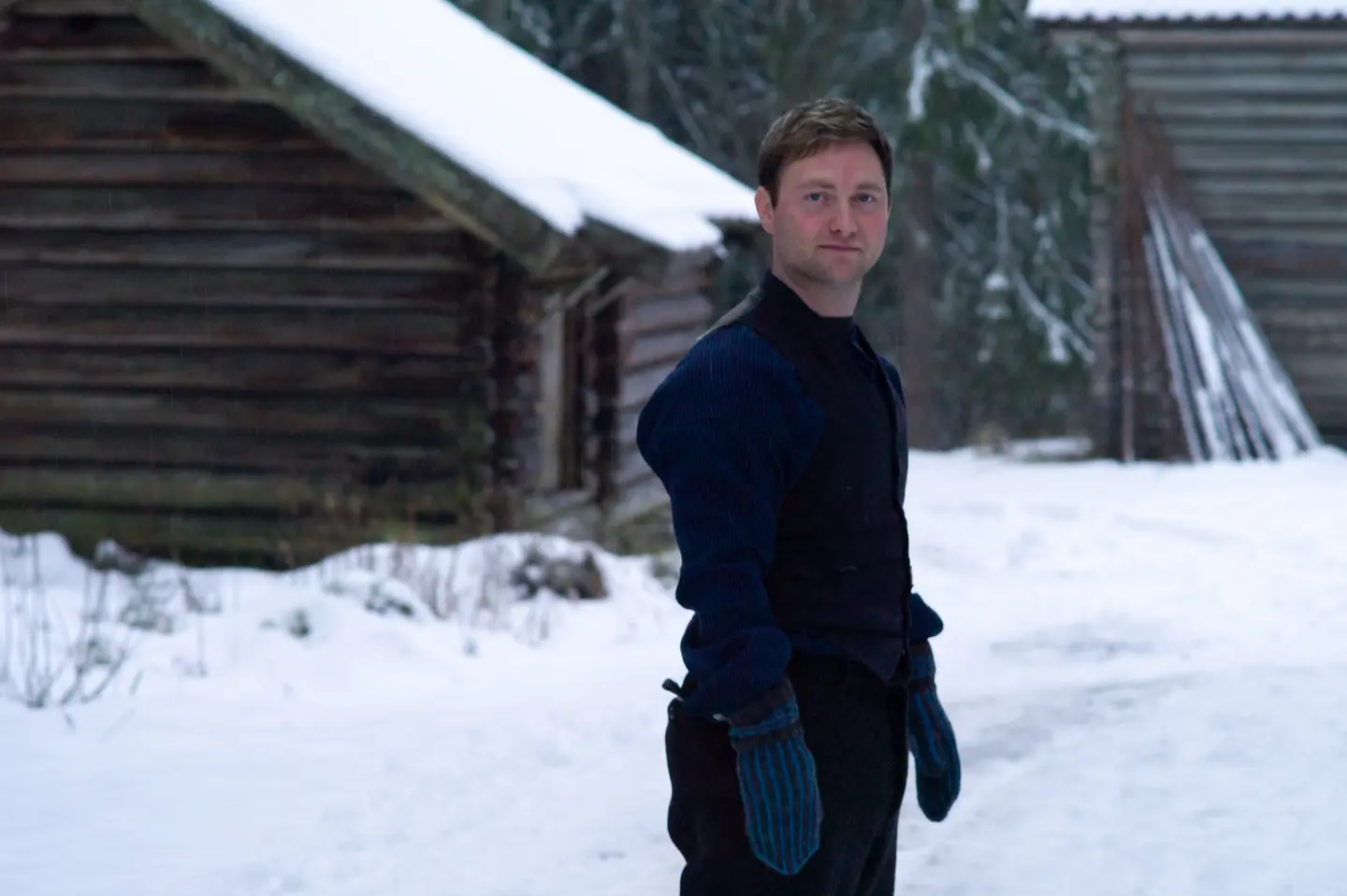 En ung gutt kledd som en dreng anno 1900 står ute på tunet. Gutten ser rett i kameraet. I bakgrunn er det to tømmerbygg. Det er snø på bakken.