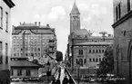 Vykort med motiv från Sandgatan i Norrköping. I bilden ses den nya spången anlagd 1926 efter skapandet av Strömsparken vid Revensgrund. I bakgrunden ses Rådhuset med Gull-Olle på sin tron överst på tornet. Där hitom på vänster sida av Nya Rådstugugatan ligger bankpalatset uppfört 1899-1902. Byggnaden till höger hitom Rådhuset är Varmbadhuset, Byggnaden vänster om bron är Klapphuset.