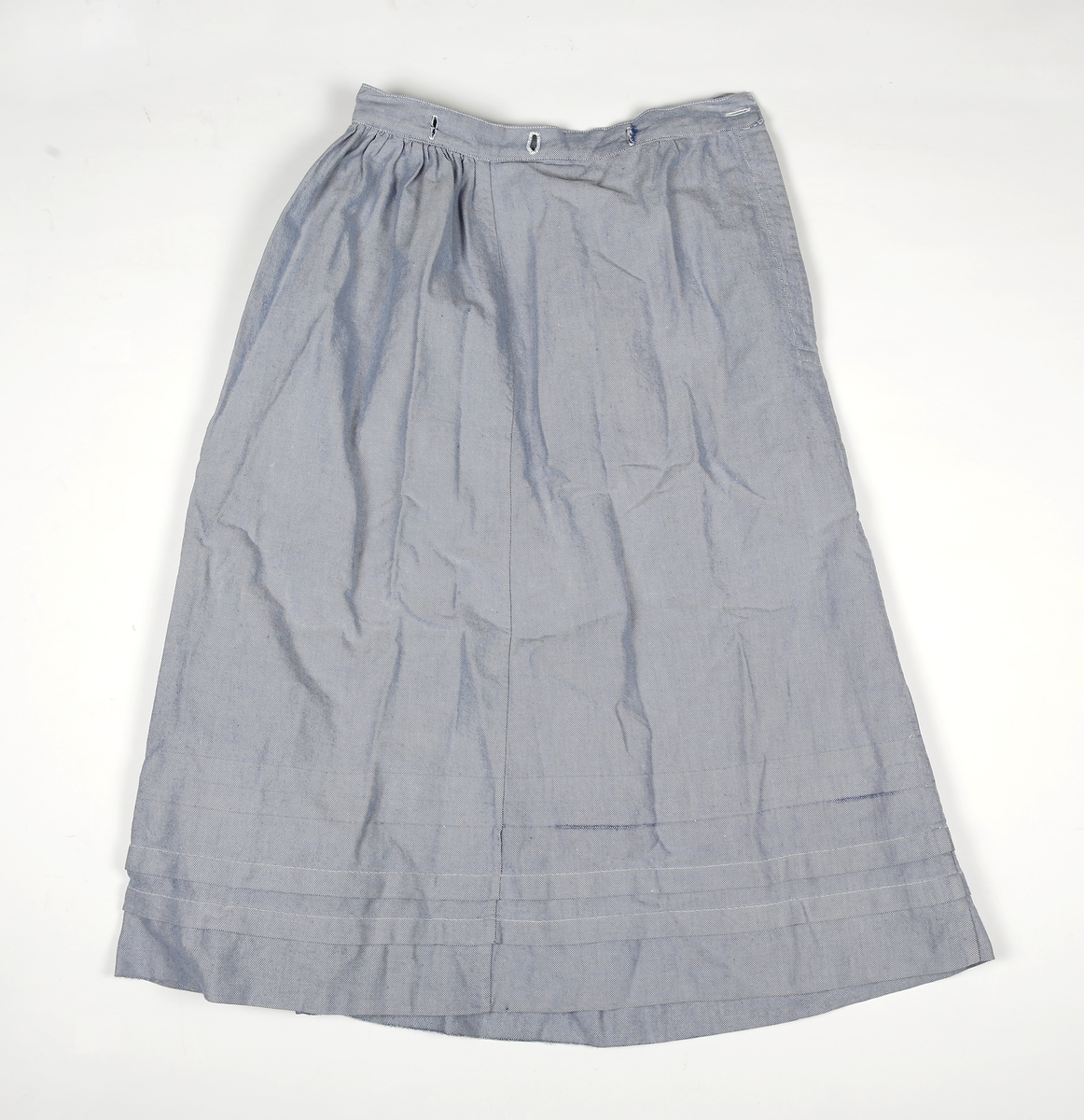Skjørt bestående av tre paneler. Skjørtet er rynket til en linning unntatt foran hvor stoffet ligger glatt. Linningen har knapphull for å kunne kneppes på blusen. Splitt i siden som lukkes med trykknapper. nederst på skjørtet er det to falder og spor etter en tredje.