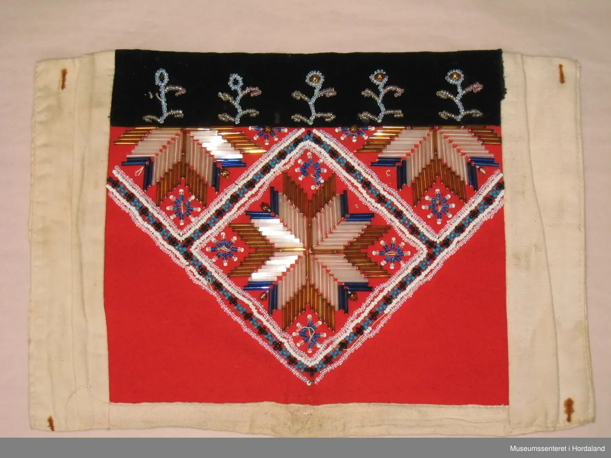 Perlebrystduk med perlebroderi sydd på raudt ullstoff. Motivet er åttebladroser med firkanta border rundt, ei heil åttebladrose på midten, ei halv oppe på kvar side. Åttebladrosene er sydd med stråperler, resten med småe perler. Store, runde, gullfarga perler til pynt innimellom, ein god del av desse er knuste. Øvst oppe er sydd på eit svart fløyelsband. Dette er pynta med fem blomstrar sydde av perler. Brystduken er montert på eit naturfarga bomullsstoff. Rustmerker etter stiftar i alle fire hjørna.