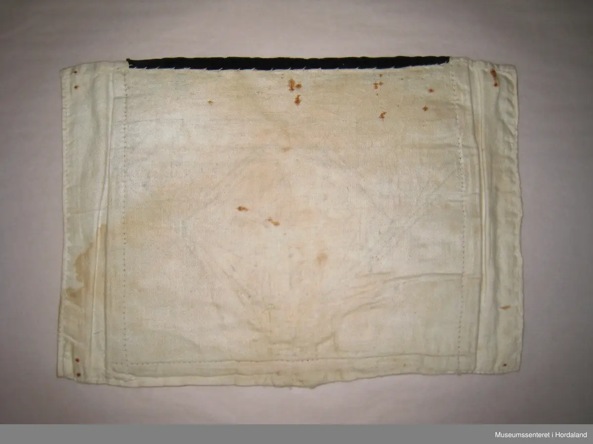 Perlebrystduk med perlebroderi sydd på raudt ullstoff. Motivet er åttebladroser med firkanta border rundt, ei heil åttebladrose på midten, ei halv oppe på kvar side. Åttebladrosene er sydd med stråperler, resten med småe perler. Store, runde, gullfarga perler til pynt innimellom, ein god del av desse er knuste. Øvst oppe er sydd på eit svart fløyelsband. Dette er pynta med fem blomstrar sydde av perler. Brystduken er montert på eit naturfarga bomullsstoff. Rustmerker etter stiftar i alle fire hjørna.