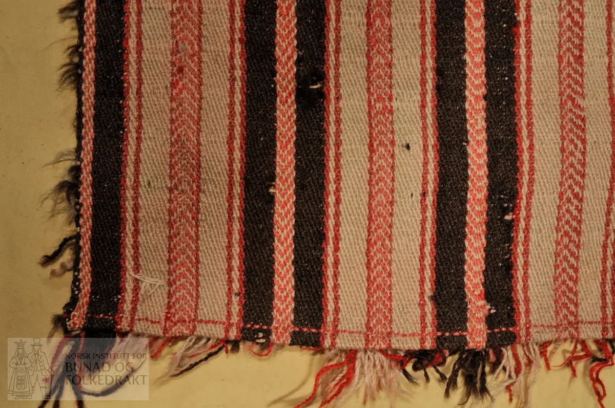 Båtrye, vevd av ull i kypert. Knyttet på floss underveis i veven. Farger i stripene /renninga : Sauesvart, rødrosa og ubleika . 	Innslaga er ubleika ull. Floss i rosa og ubleika ull.