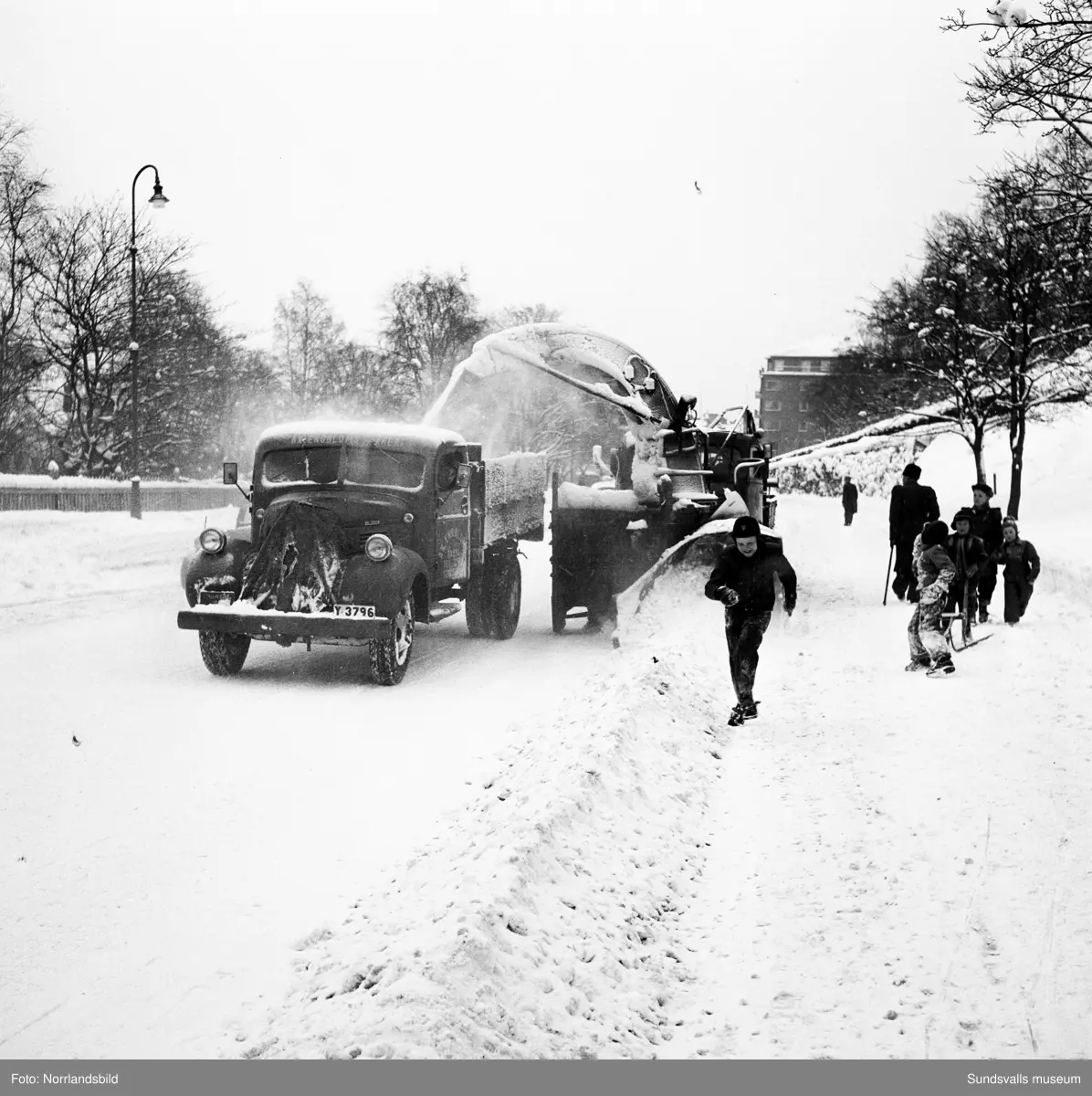 Snöröjning med plog, lastbilar och snöslunga på västra Storgatan. Några barn leker en farlig lek framför snöslungan.