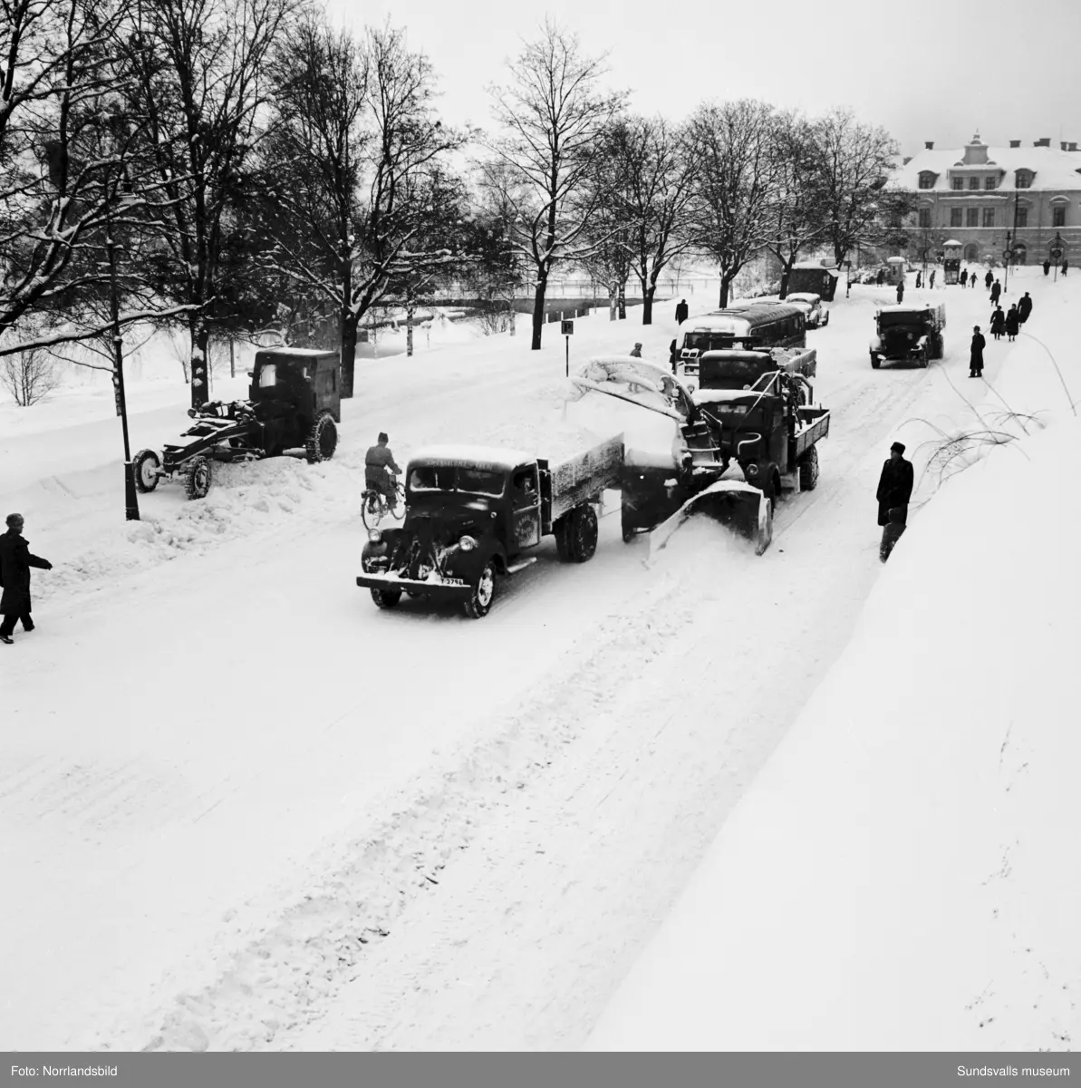 Snöröjning med plog, lastbilar och snöslunga på västra Storgatan. Några barn leker en farlig lek framför snöslungan.