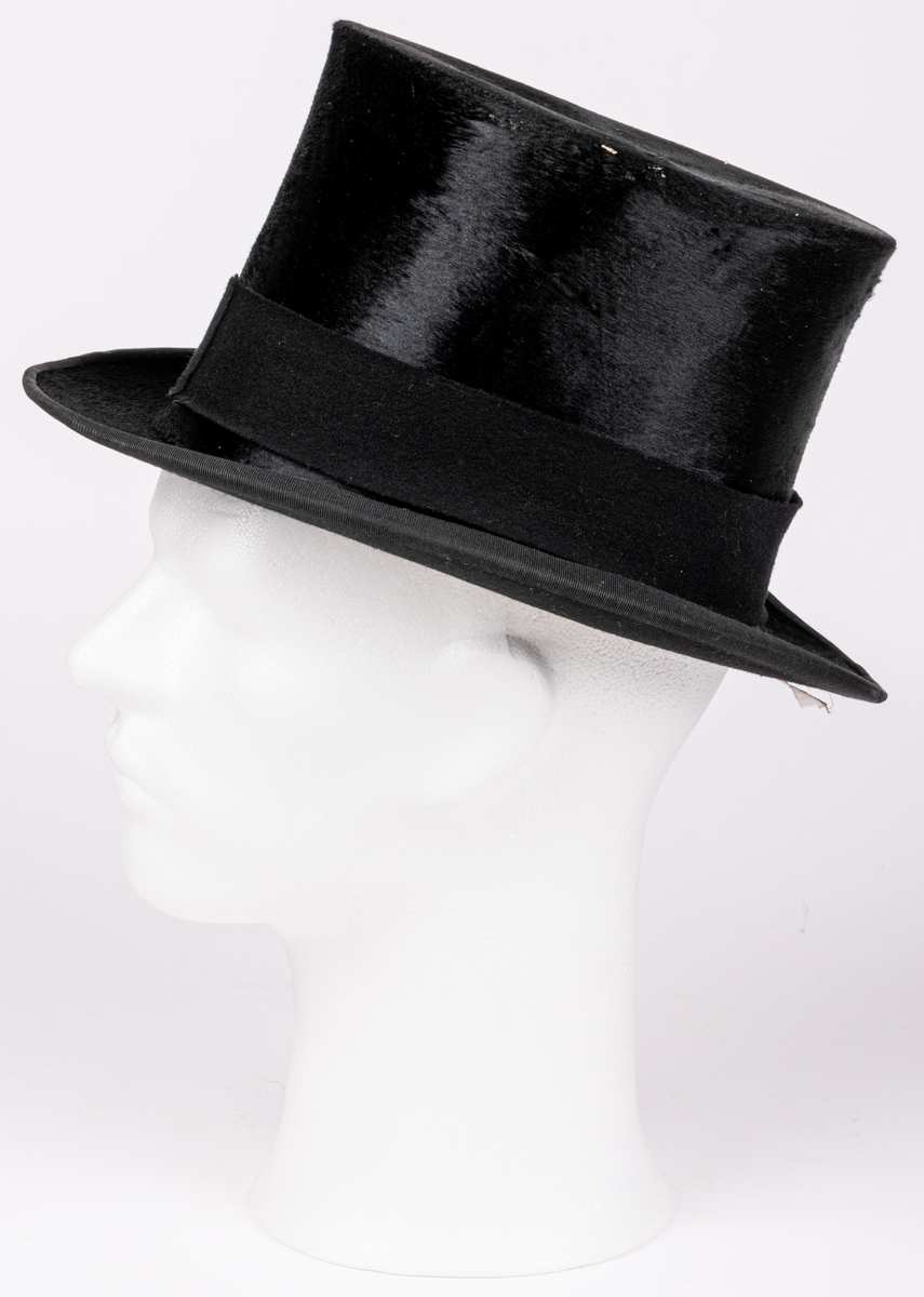 Stormhatt märkt Aktiebolaget J.W Haglund, N. Kungsgatan 1, Gefle. Svart.
Höjd 14cm, brättbredd 4,5cm.
Hattask av svart papp med guldtext på locket. English manufacture of hats. Best London deposed.