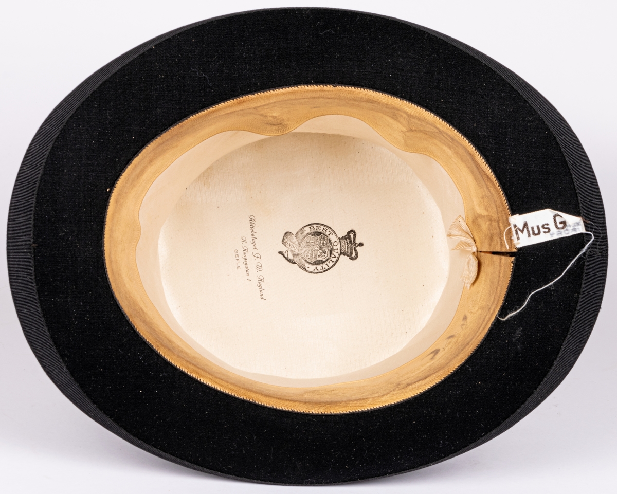 Stormhatt märkt Aktiebolaget J.W Haglund, N. Kungsgatan 1, Gefle. Svart.
Höjd 14cm, brättbredd 4,5cm.
Hattask av svart papp med guldtext på locket. English manufacture of hats. Best London deposed.