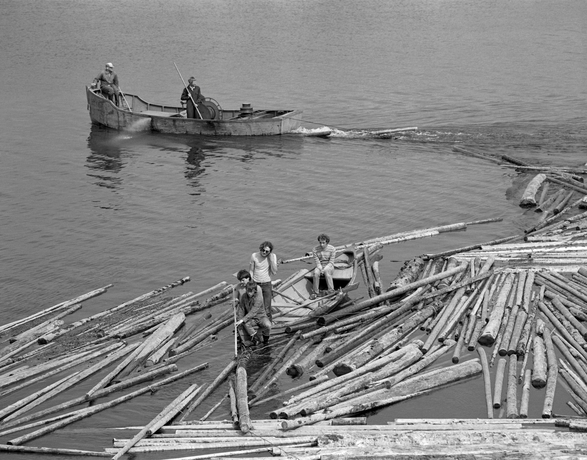 Løsning av tømmerslep ved Sleppetangen i Spydeberg i Østfold, der nedre del av Glomma renner ut av innsjøen Øyeren.  Fotografiet later til å være tatt fra et høyt punkt, antakelig en bergknau, ned mot fløterne,  I forgrunnen arbeider fire unge menn fra en robåt.  To-tre karer i stevnen av båten arbeider med en fløterhake, antakelig for å løsne en knute på vaieren som under foregående sleping fra Fetsund og sørover innsjøen har holdt «mosene» (tømmerbuntene) i slepet samlet.  Fjerdemann sitter påtofta i den andre enden av båten.  Her ved Sleppetangen ble både slepene og de enkelte mosene ble løst opp, slik at stokkene kunne flyte løst gjennom tømmerrennene forbi kraftverkene i elvepartiet nedenfor.  I bakgrunnen på dette fotografiet ser vi en større og mer robust motordrevet jernbåt, «Kari», som ellers ble mye brukt i «etterrensken».  Arbeidet som vises på dette fotografiet ble altså utført fordi virket måtte fløtes løst øverst i nedre Glomma, hvor stokkene skulle gjennom tømmerrennene ved kraftverkene Solbergfoss, Kykkelsrud og Vamma, før de igjen kunne samles, sorteres, moses og slepes videre fra Glennetangen lense i Skiptvet. 