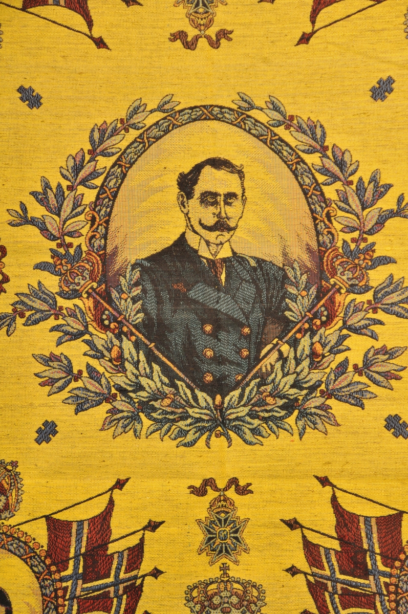 Tekstil i bomull med litt metalltråd. Fleire fargar. Portrett av HaakonVII på midten. I kvart hjørne er der portrett av Kong Haakon, Dronning Maud og Kronprins Olav. Den norkse løva med teksten "Haakon VII Norges Konge", Flagg, ranker og løver m.m. Tafs kring alle kantane. Tafset er ein forlengelse av varp og verft. Tydeleg rett og vrangside. God stand, men noko bleika på den eine sida.
