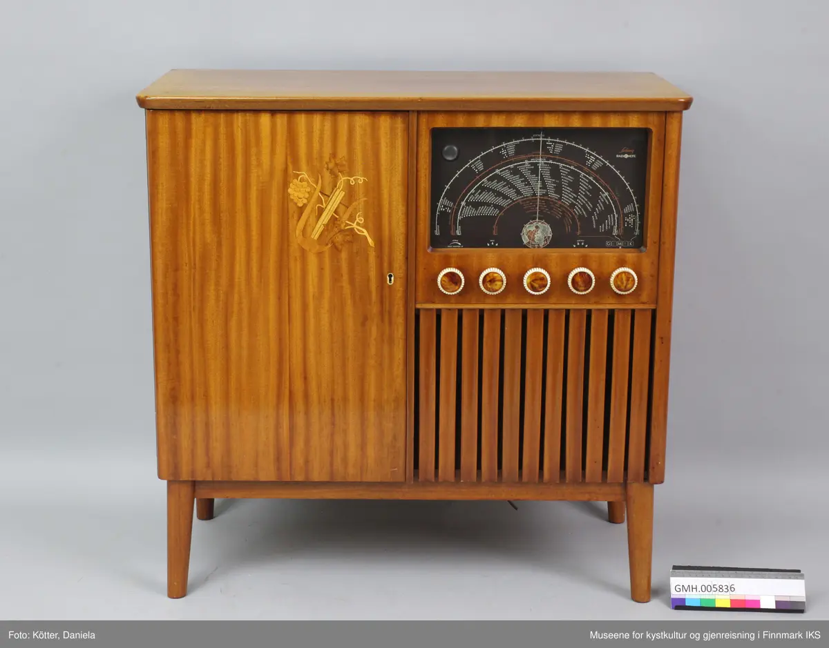 Radiokabinettet er laget av finert og transparent lakkert tre. Korpuset hviler på ei ramme som beinene er festet til. Beina er dreide, smalner nedover og skrår lett utover. Fronten er inndelt i to. Radiodelene befinner seg til høyre, med med oversikten over kanaler for FM, kortbølge og langbølge oppe, fem runde knapper i midten og plass til høyttaleren nede som mangler. Høyttaleren skjules av vertikale pyntelister og porøs stoff bak disse. På venstre siden av skapet er det en dør med et intarsiebilde av en lyre, et type harpe, og en vinranke. Bak den ligger det to fag. Den øverste er til et grammofon, den nedre til oppbevaring av plater. Døra kan låses og nøkkelen følger med. Bare er den så deformert, at den ikke passer inn i låsen og døra forbli dermed lukket. Bakveggen av radiokabinettet mangler.