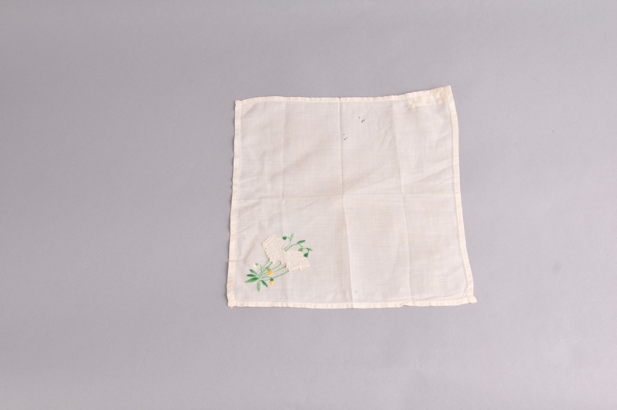 Kvadratiske lommetørkle i tynnt bommullstøy. Blomsterbroderi og applikerte blomar nede i eine hjørnet.Smal holfald kring kanten. Lommetørkledet er brodert med maskin.
