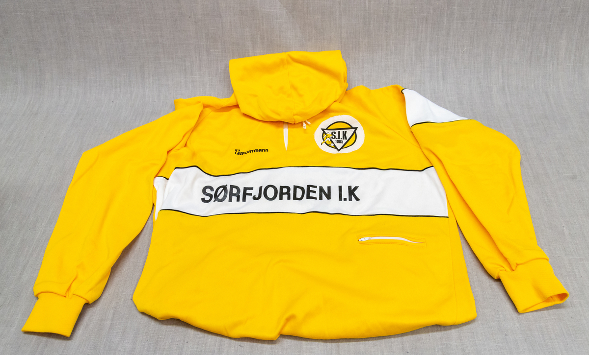 Hettegensar med gul hovedfarge og kvit effektfarge. Kort, kvit glidelås i front og i brystlomme på venstre side. Kvit snor i hette. 