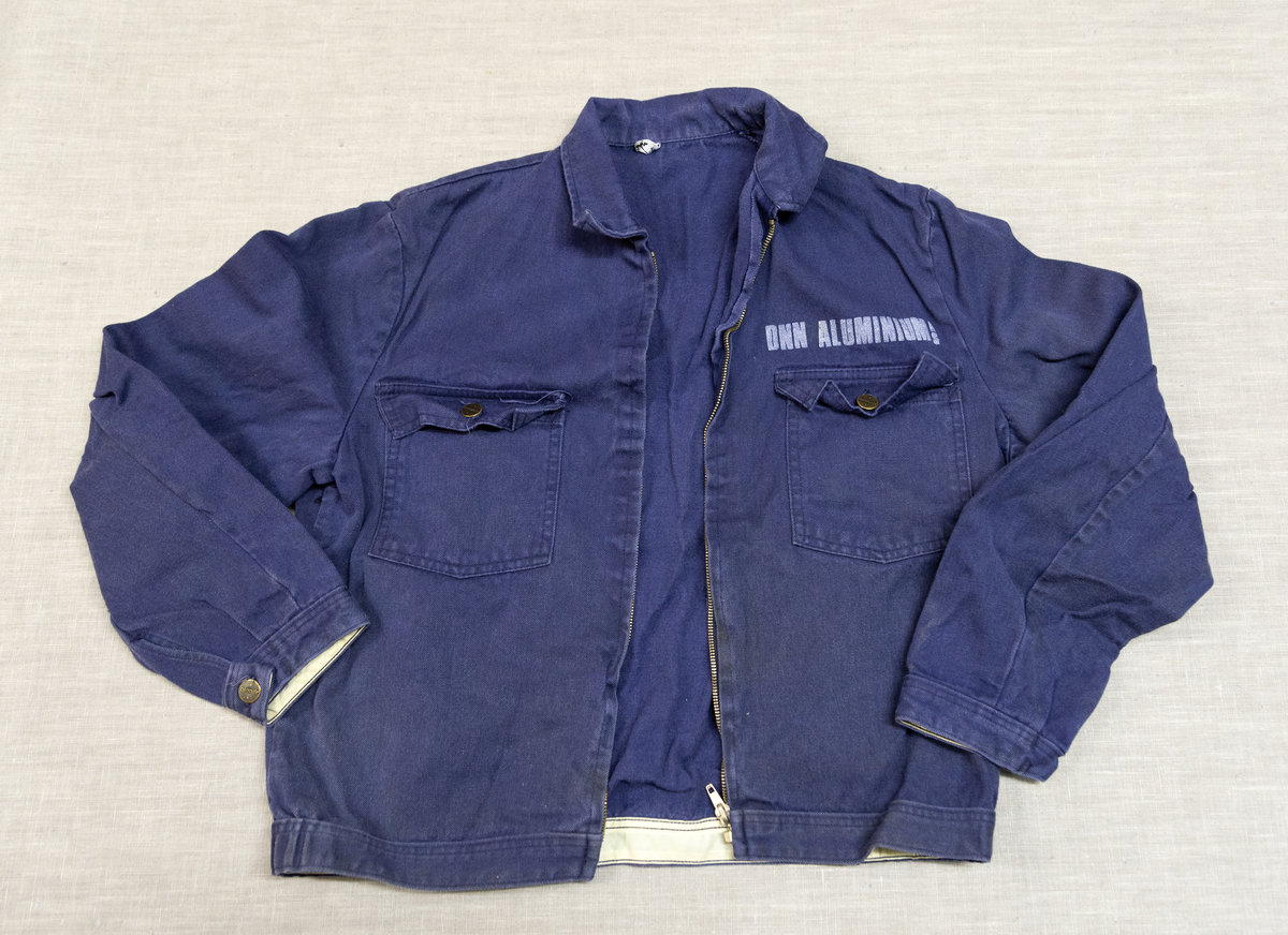 Todelt dress i mørk blå bomullsdenim. Storleik 48 (båe delene). Glidelås i metall framme i jakke, knepping med metallknappar i buksegylf. To brystlommer på jakka, to stikklommer i buksa. Innside av bukselinning i tynt nylonstoff (?). Buksa har produsentmerket laminert fast i høgre lomme og jakka har tilsvarande på innsida bak høgre brystlomme. Vaskeanvisning 60 gr. 

Buksa har fleire større og mindre hol, særleg nede på venstre buksebein, - både framme og bak. Om desse er forårsaka av varme eller syre er ikkje kartlagt, men fargen tyder på høg varme. Det er ikkje gjort forsøk på reparasjon av hola. Stoffet er ikkje flammehemma.