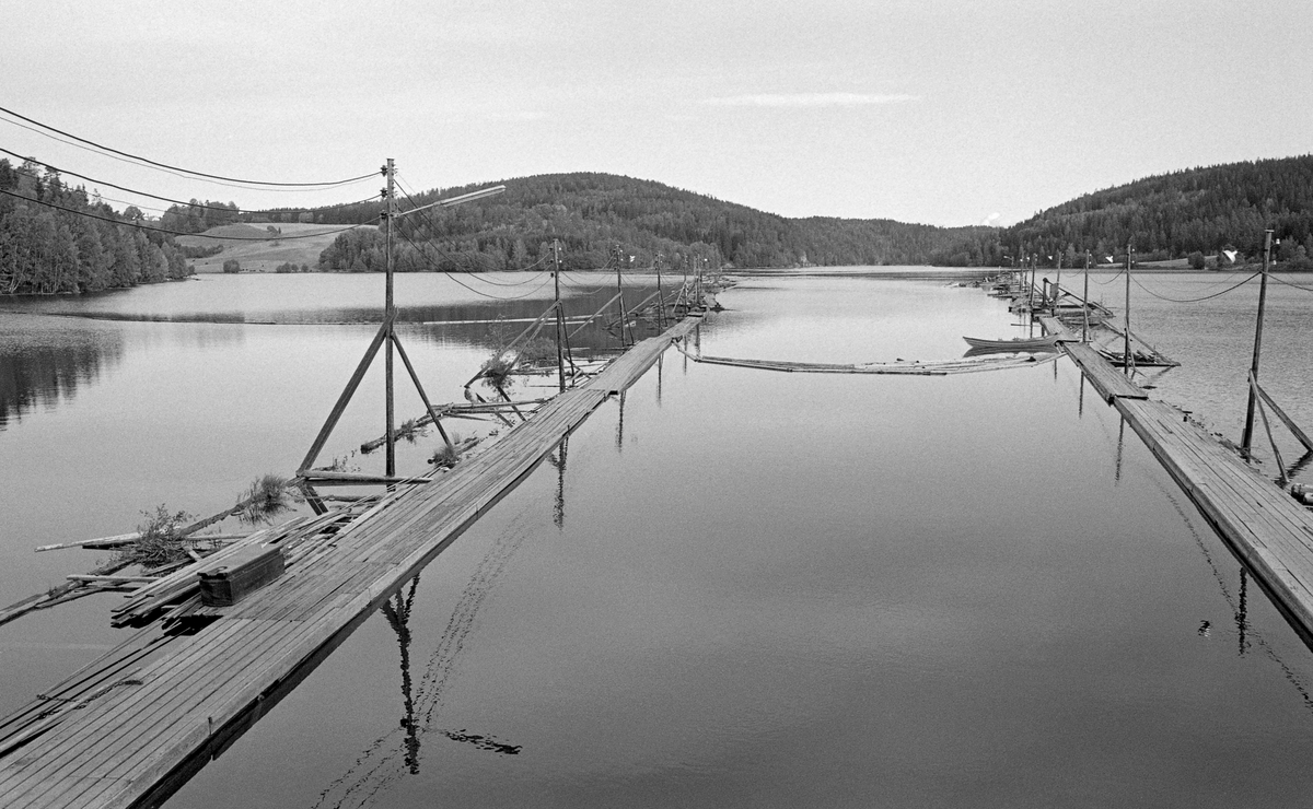 Innløpet til tømmertunnelen forbi dammen ved Solbergfoss kraftstasjon i nedre Glomma, der elva er grense mellom kommunene Spydeberg og Askim.  Tømmertunnelen ble sprengt inn i fjellet på vestsida (Spydeberg-sida) av elveløpet.  Dette fotografiet er tatt fra en posisjon over tunnelinnløpet.  Vi ser lensa som skulle lede tømmeret mot tunnelen, med gangbruer på begge sider, der fløterne kunne gå med haker og sørge for at stokkene gikk riktig mot tunnelen.  Fotografiet viser at den indre delen av lensa hadde lyktestolper, noe som gjorde det mulig å kjøre kveldsskift her utover høsten, når kveldene begynte å bli mørke.  I bakgrunnen til venstre ligger Tonnerudvika, mens Glomma kommer ned i en forsenkning i det skogkledde terrenget, rett opp for inntakslensa midtakse. 