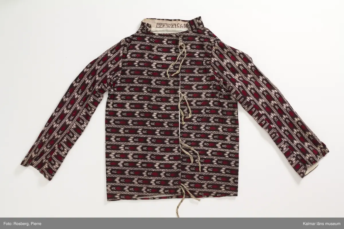 KLM 35481 Tröja. Sydd av kattuntryckt bomullstyg, svart botten med tvärgående ränder i bomullstygets bottenfärg samt i mörkrött. Tröjan är skuren i ett stycke utan sidsömmar. Lång ärm hopsydd av två delar med längsgående sömmar, den ena delen är skuren på längden den andra delen är skuren på tvären. Sprund vid handlederna, rund halsringning med ståkrage. Knyts framtill med gråvitrandiga band av linne. Fodrad med vitt bomullstyg. Halskantens foder av tygets avslutningskant med tryckt text: Blecktornet 1808 95 AB Möller. Tyget är tryckt på Blecktornets Kattuntryckeri på Södermalm, Stockholm.