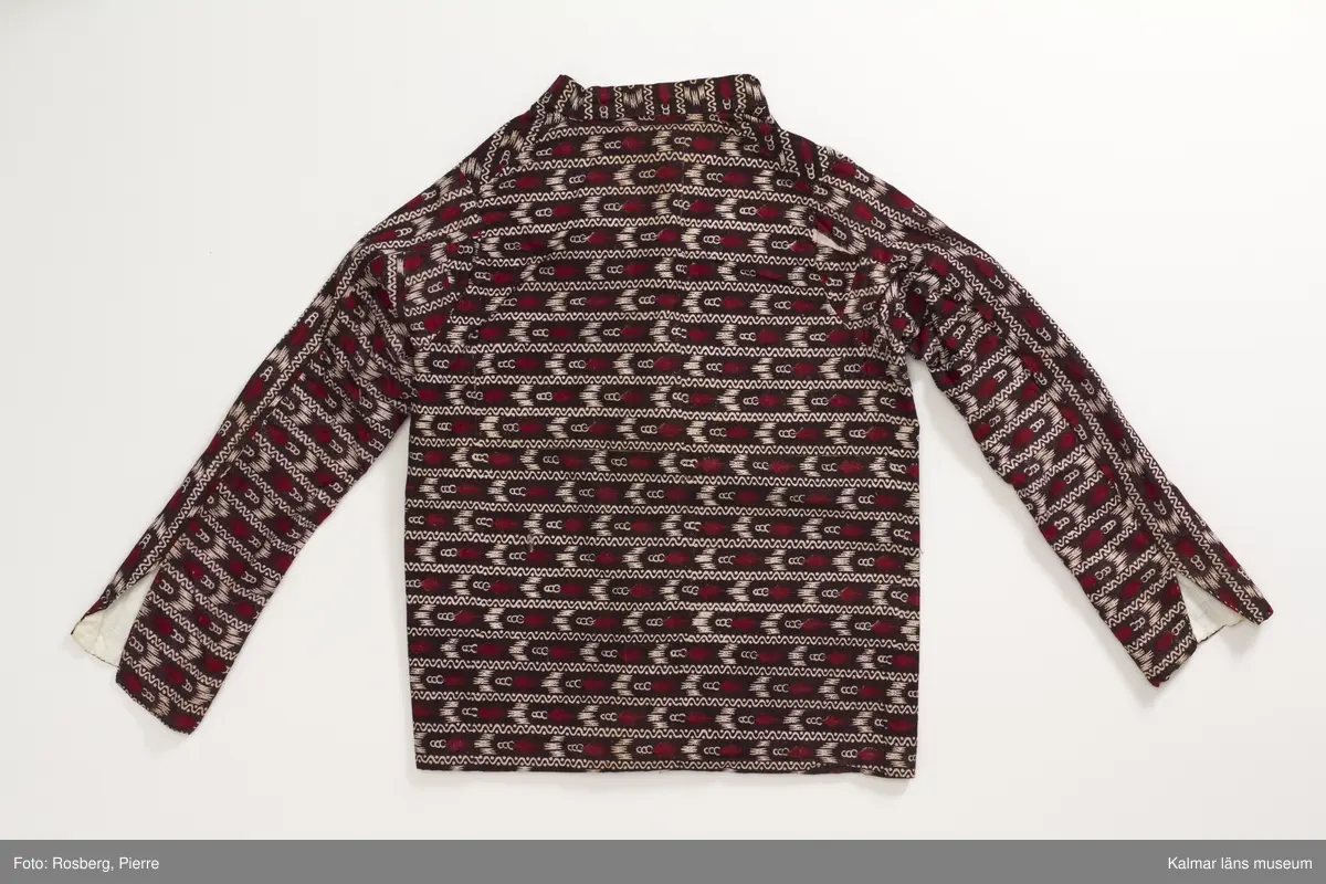 KLM 35481 Tröja. Sydd av kattuntryckt bomullstyg, svart botten med tvärgående ränder i bomullstygets bottenfärg samt i mörkrött. Tröjan är skuren i ett stycke utan sidsömmar. Lång ärm hopsydd av två delar med längsgående sömmar, den ena delen är skuren på längden den andra delen är skuren på tvären. Sprund vid handlederna, rund halsringning med ståkrage. Knyts framtill med gråvitrandiga band av linne. Fodrad med vitt bomullstyg. Halskantens foder av tygets avslutningskant med tryckt text: Blecktornet 1808 95 AB Möller. Tyget är tryckt på Blecktornets Kattuntryckeri på Södermalm, Stockholm.