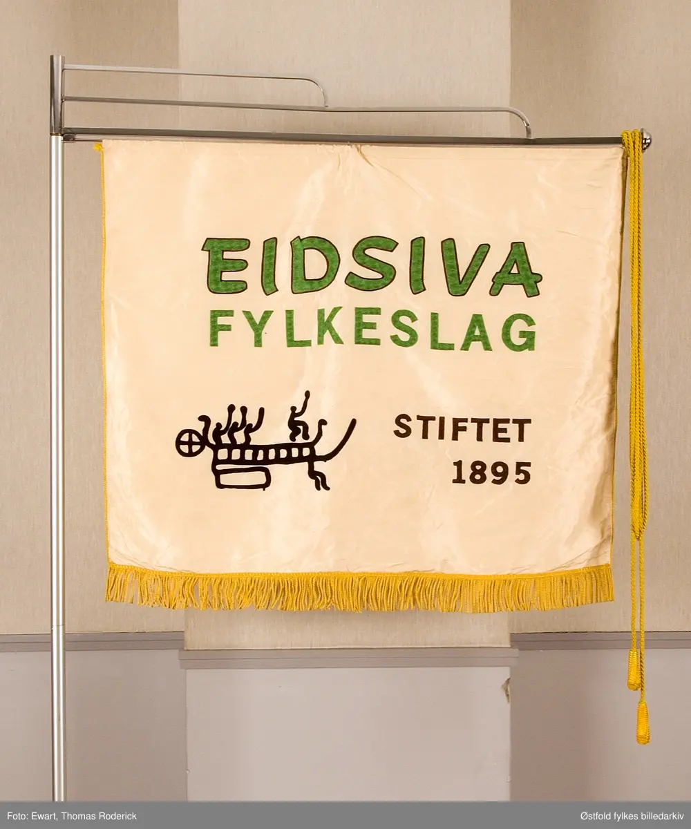 Fane til Eidsiva fylkeslag, stifta 5. januar 1895 under navnet Eidsiva Ungdomslag., fylkeslag i NU. Maskinbrodert banner 120 x 100 cm frå ca. 1979. Motiv: kopi av ei av helleristningane i Skjeberg. Teikna av Bjørn Hansen.
Adresse: Sverres gate 4. Fredrikstad.