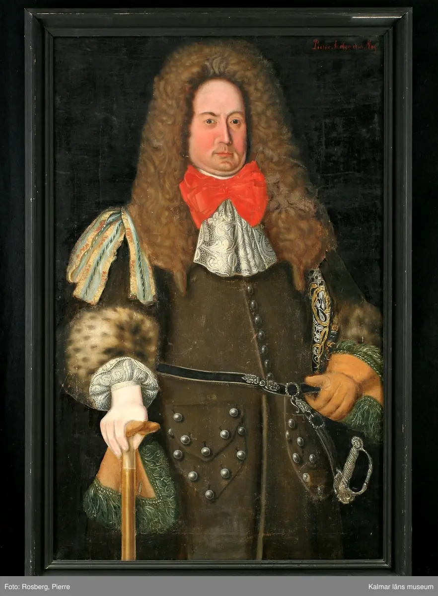 Porträtt av Magnus Gabriel Craelius, Fortifications-Capitain i Kalmar. Grön livrock av polskt snitt. Skinnuppslag på ärmarna. Stora ficklock med många knappar. Värja och gehäng. Spetskrås vid hals mm. Peruk. I högra handen käpp och handske, på vänstra handen älghudshandske.