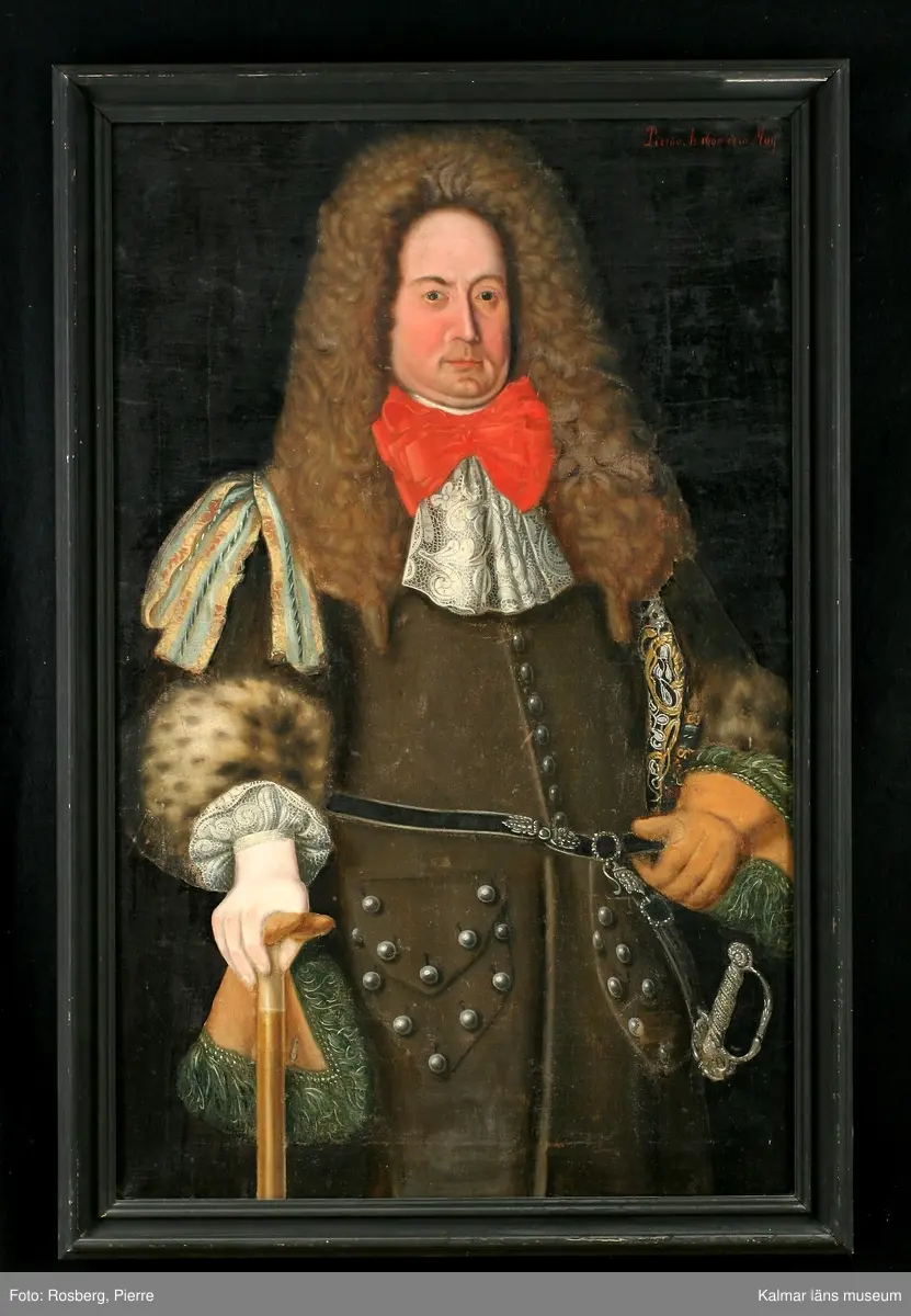 Porträtt av Magnus Gabriel Craelius, Fortifications-Capitain i Kalmar. Grön livrock av polskt snitt. Skinnuppslag på ärmarna. Stora ficklock med många knappar. Värja och gehäng. Spetskrås vid hals mm. Peruk. I högra handen käpp och handske, på vänstra handen älghudshandske.