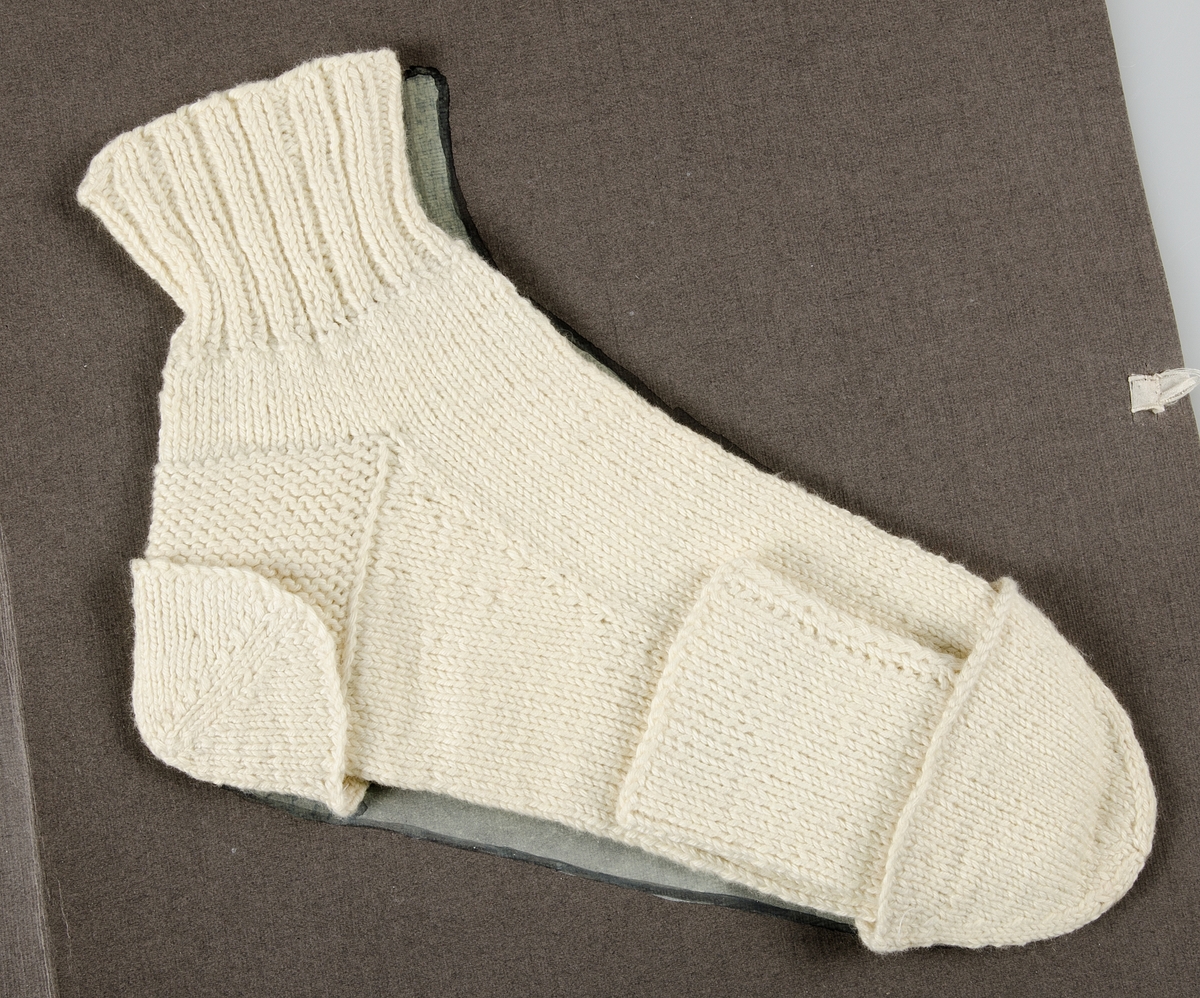 Stickad socka av oblekt bomullsgarn, samt tre stickade delar till strumpfot av oblekt bomullsgarn. Förvaras i en grå papperspärm, på framsidan texten: STRUMPFOT JUDIT JANSSON. På uppslaget en målad socka. Knytes med ljusa silkesband.