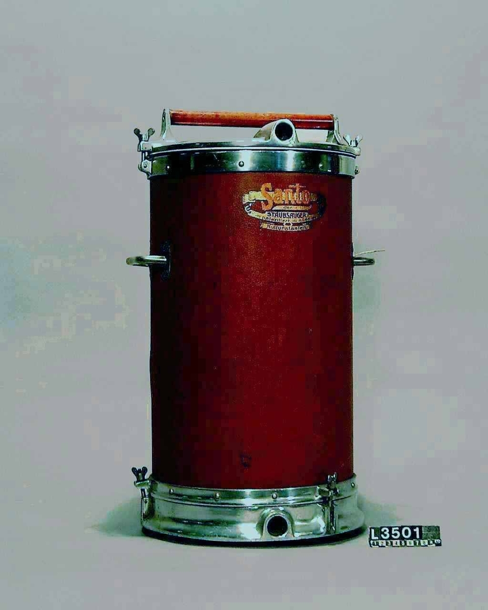 Stående cylindrisk dammsugare. Topplock av metall och cylinder, klädd med röd-brun pegamoid. Dammsugaren väger ca. 20 kg och är utrustad med både motor och pump.