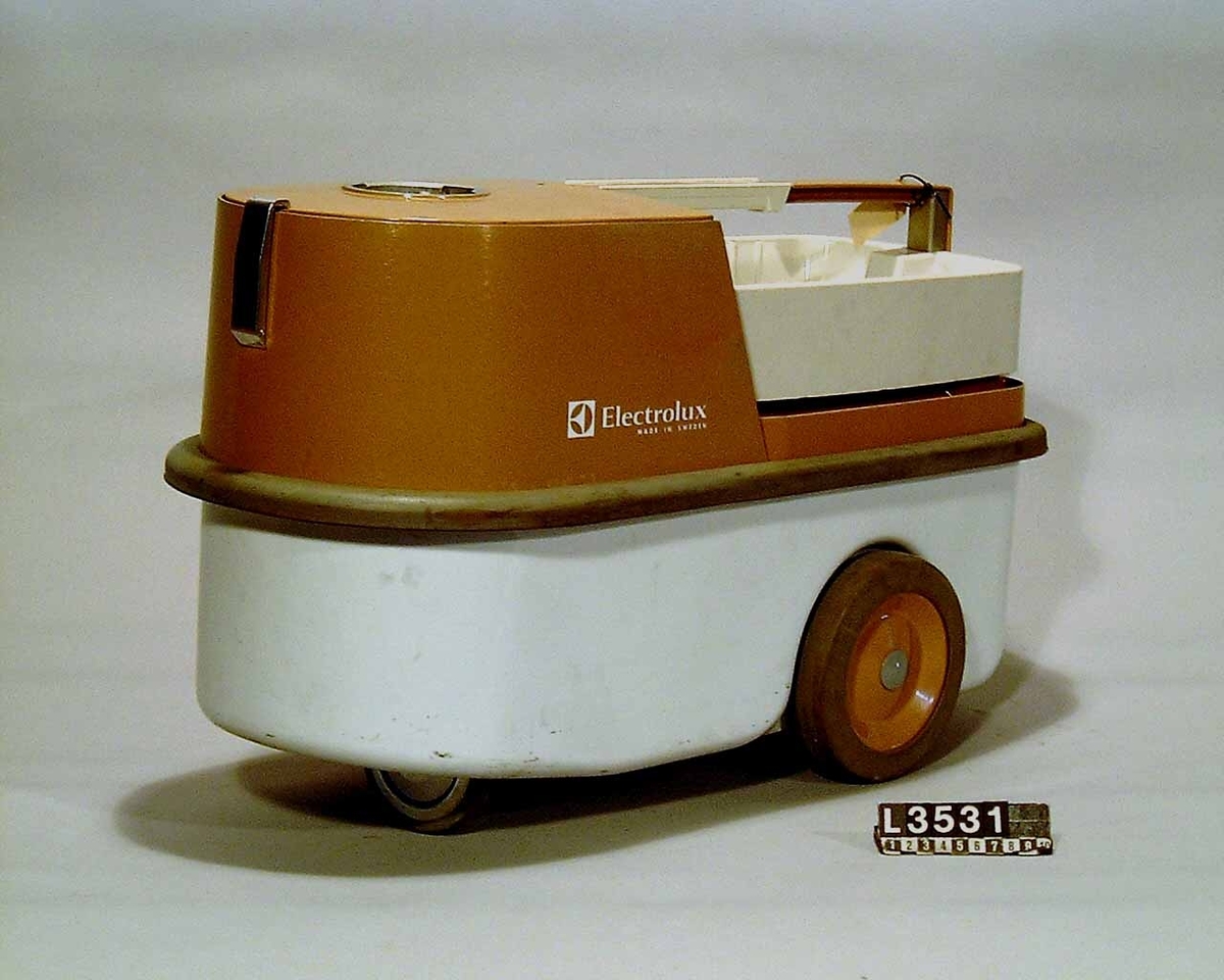 Industri-/hotell- dammsugare med två motorer. Dammsugaren har två strörre hjul på  sidorna och ett mindre styrhjul fram. Apparaten är färgsatt i brunt och vitt.