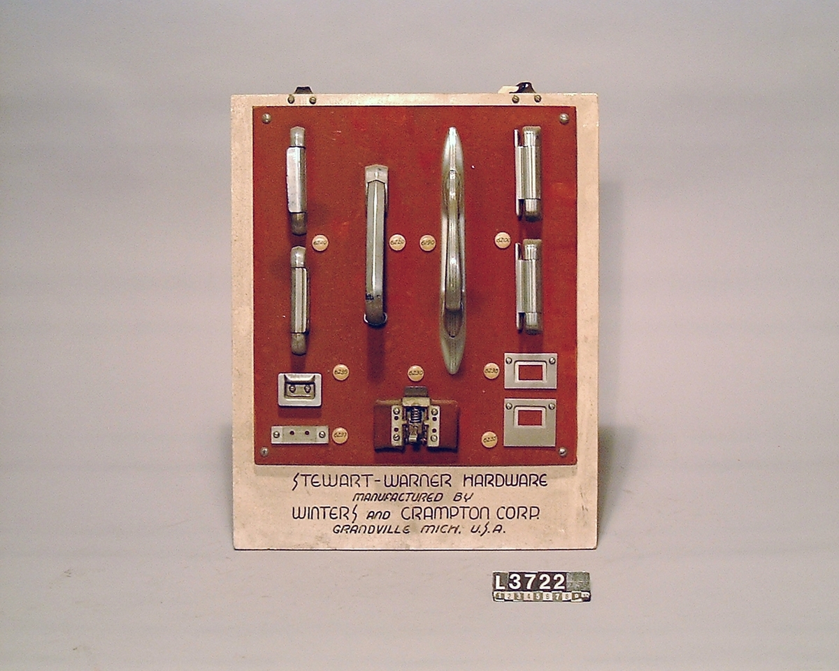 Gångjärn, handtag och låsannordningar till Stewart-Warner kylskåp. Samtliga delar är uppsatta på en träplatta som är klädd med tyg. Märkt: Sewart-Warner hardware.