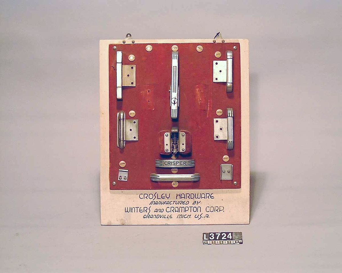 Gångjärn, handtag och låsannordningar till Crosley kylskåp. Samtliga delar är uppsatta på en träplatta som är klädd med tyg. Märkt: Crosley Hardware.