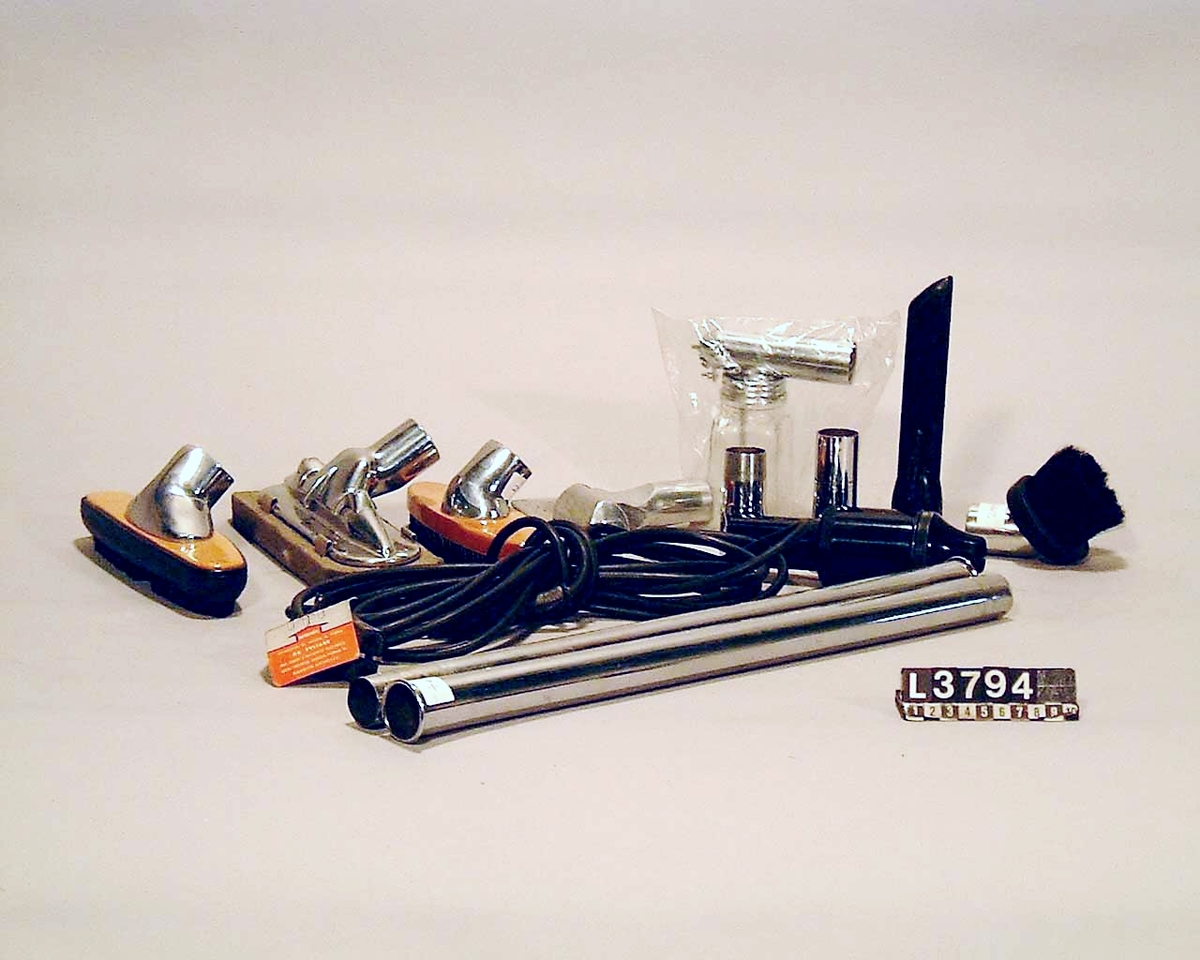 Cylinderdammsugare i grön metallic med fram och bakstycke i blank metall. Medar i krom med hjul under. Kabel, rör, munstycken och vätskespridare finns. Apparaten med tillbehör ligger i en koffert/väska. Slangen saknas.