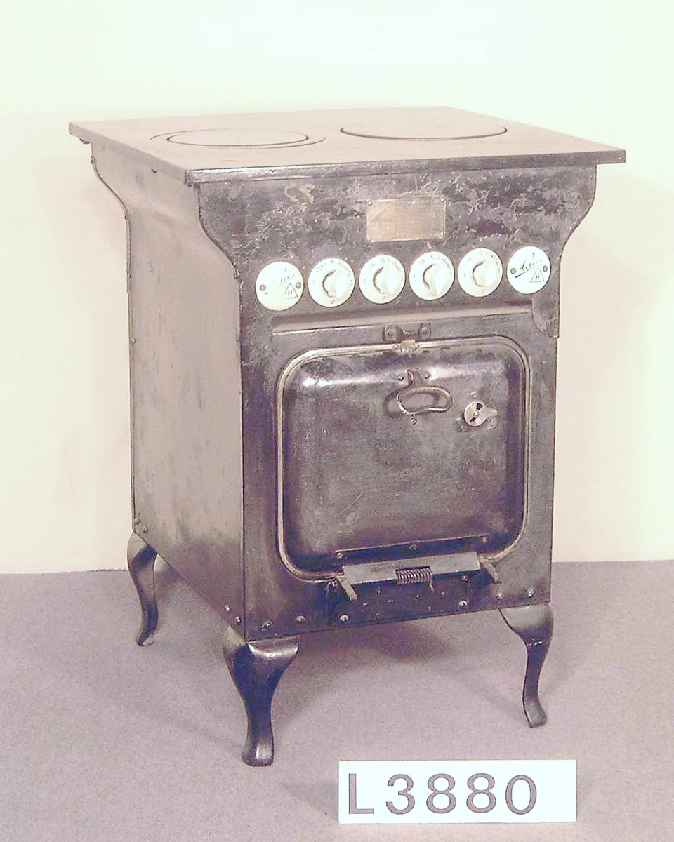 Elektrisk spis i svartmålad plåt och gjutjärn. Två st. kokplattor med effekten 850 & 1200 W. För ugnen finns två strömbrytare för över och undervärme. Fyra st. vita porslinsströmbrytare för plattor och ugn. På vardera sidan om strömbrytarna sitter emaljerade skyltar med Helios på. Överhällen är uppfällbar. Gjutjärnshandtag på ugnsluckan. Brickan med Elektrolux inventarienr. sitter under överhällen.