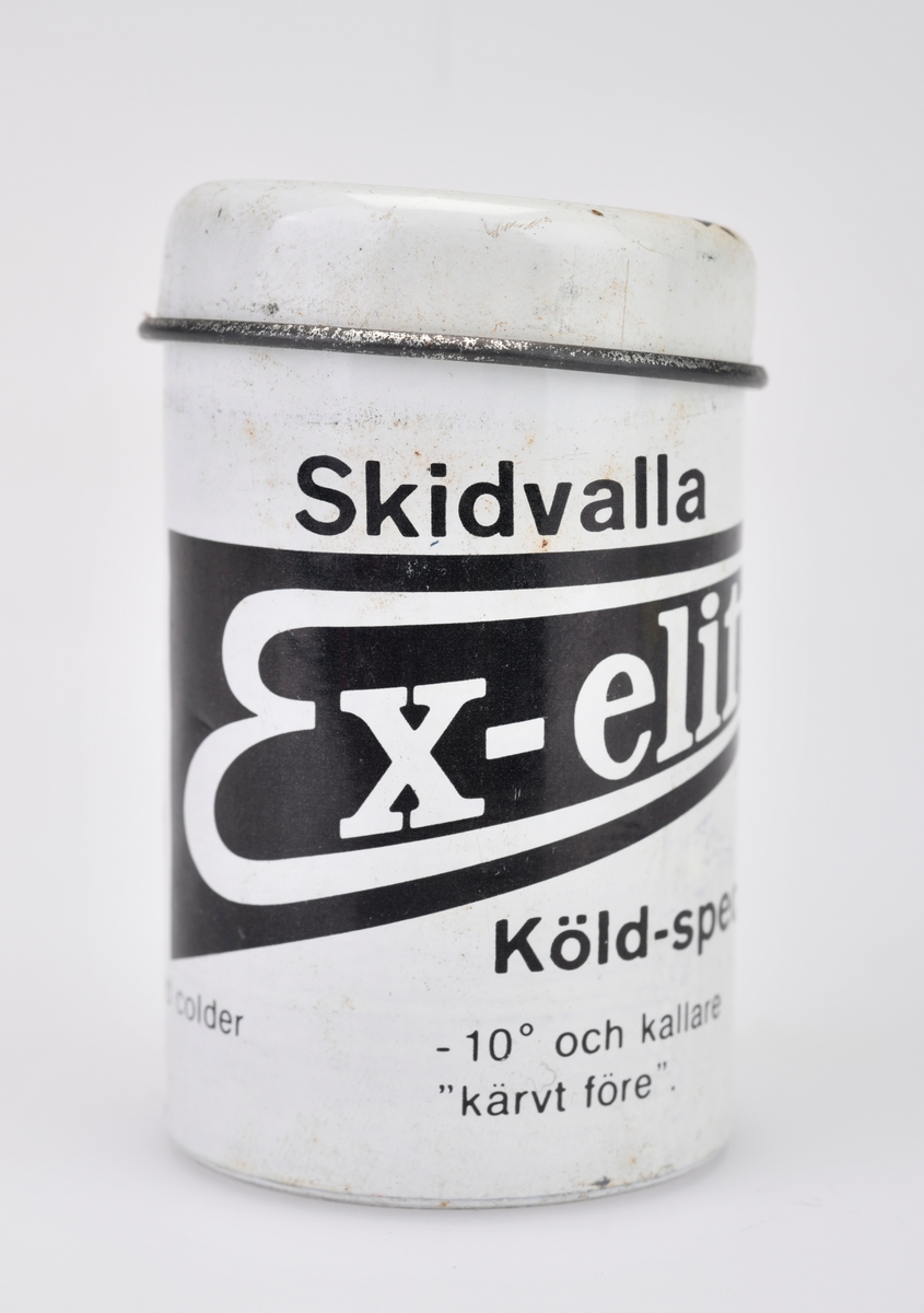 En boks/sylinder med skismurning av typen Ex-elit "Köld-special". Boks og lokk er av metall. Metallet skal rives av etter hvert som det brukes av skismurningen. Boksen er hvit og svart med svart og hvit skrift, lokket er hvitt med et svart felt med hvit skrift. For tekst på for- og bakside - se "Påført tekst/merker".  Det er informasjon om bruk på svensk, engelsk og finsk. Voksen er svart på farge.