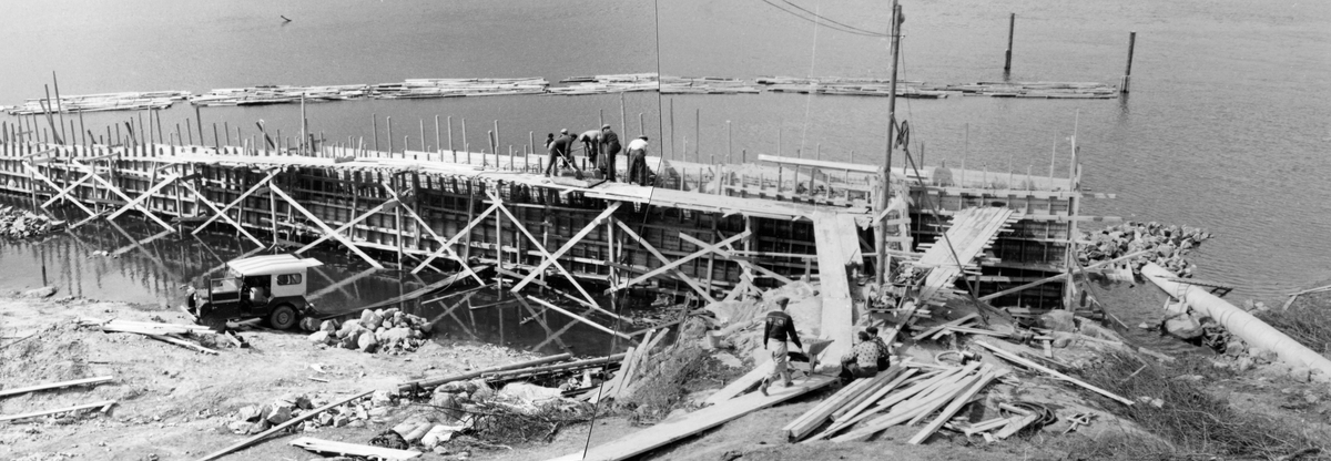 Fotomontasje fra arbeidet med å støpe kai til Glomma fellesfløtingsforenings båter i den nedre delen av vassdraget ved Furuholmen i Varteig våren 1953.  Fotografiet er tatt fra bakken ovenfor anleggsstedet.  Vi ser en mann som kjører ei trillebår med betong på en bordgang mot forskalinga til den lange, kraftige muren.  Fra en plattform på innsida av forskalinga arbeidet fire-fem mann med spader på det punktet der det akkurat da fotografiet ble tatt foregikk støpearbeid.  Ved vannkanten, på innsida av forskalinga sto det en Jeep-bil.  I Glomma, like utenfor det som skulle bli kaimur, lå det en kjede av ”moser” (tømmerbunter) forankret i en trepæle.  Sammenlikner en denne montasjen med opptak som ble gjort et par uker tidligere (jfr.  SJF. 1990-04012), er det tydelig at vannstanden var i denne delen av Glomma var i ferd med å stige som følge av vårflommen, som sikkert innebar at anleggsarbeidet var underlagt et tidspress.  Dette er kommentert i en undertekst til fotografiet i Glomma fellesfløtingsforenings album nr. 50: ”Vannet steget over fangdammen.  Støpingen kommet opp i sikker høyde. ”