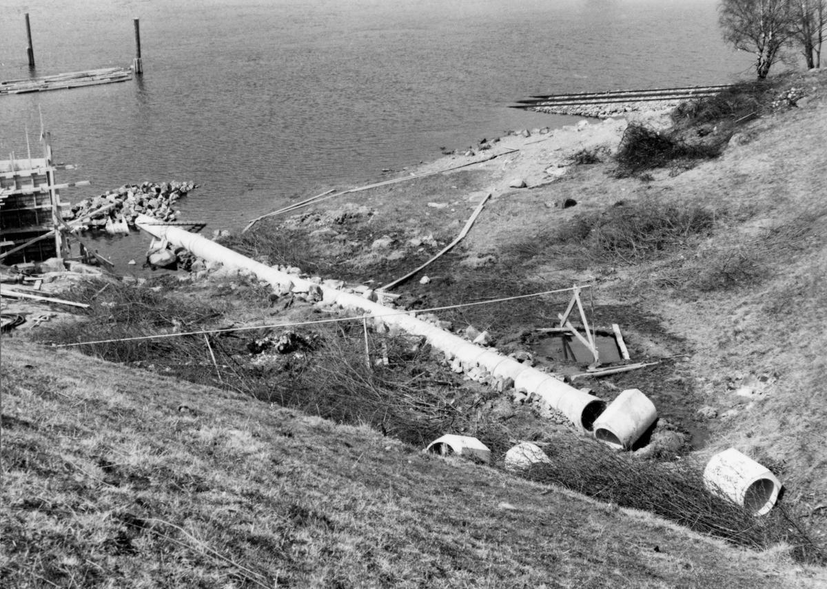 Delvis rørlagt bekkefar ved Furuholmen i Varteig, der Glomma fellesfløtingsforening våren 1953 fikk støpt en kaimur for organisasjonens båter i den nedre delen av vassdraget.  Vi ser rørgata, som ennå ikke var ferdig, sentralt i bildet, og litt av forskalinga til den nevnte muren til venstre i bildet.  Helt til høyre i bildet ser vi hvordan skinnegangen til båtslippen på Furuholmen kom opp fra vannet.  På tvers av den nevnte rørledningen, sentralt i bildeflata, ser vi en spinkel konstruksjon som bærs av lekter fra en vannpytt ved siden av rørgata.  Det er sannsynligvis denne konstruksjonen det refereres til i underteksten til bildet i Glomma fellesfløtingsforenings fotoalbum nr. 50: ”Vannledning til betongblanderen”. 