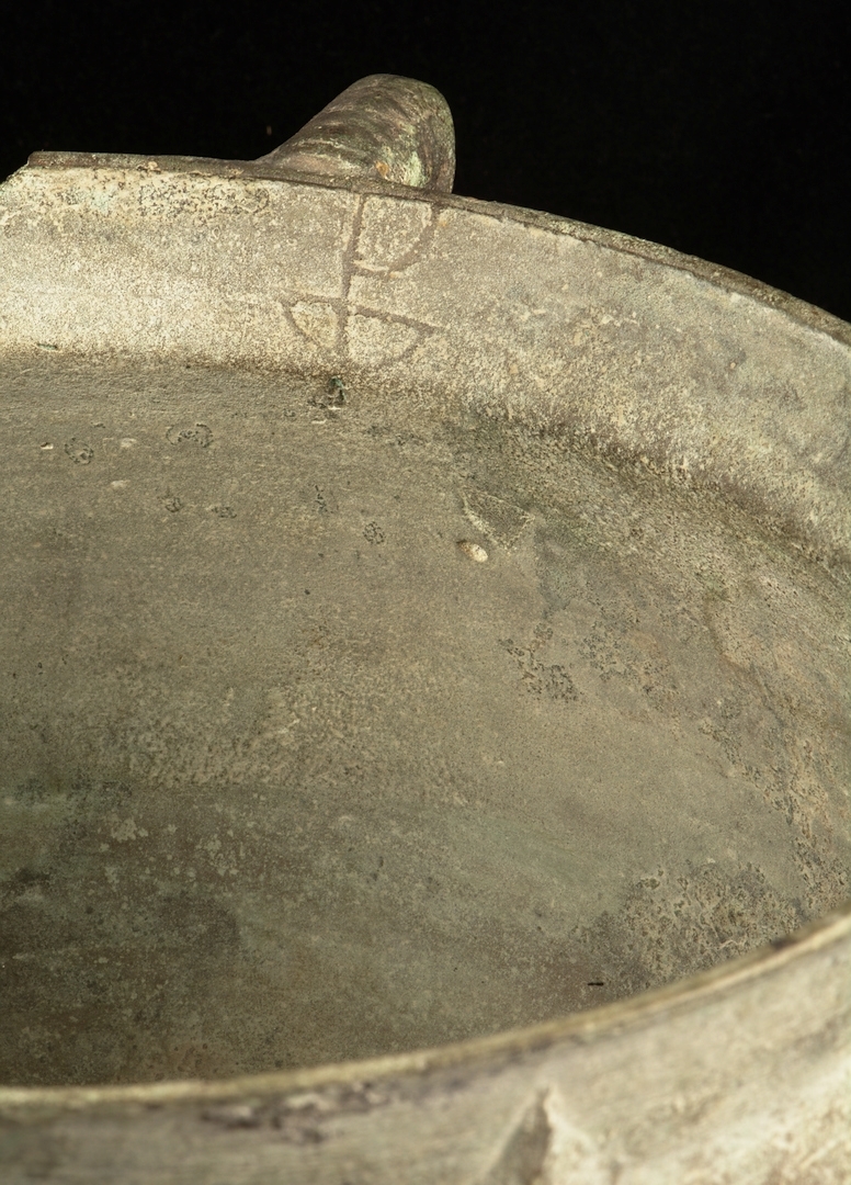 Trefotsgryta av brons. Signerad av tillverkaren i form av att upphöjt bomärke i metallen. Grytan har även en omsorgsfullt utförd lagning.

 Mynningen på grytan har en diameter på ca 25cm.