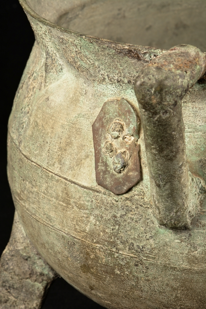 Trefotsgryta av brons. Signerad av tillverkaren i form av att upphöjt bomärke i metallen. Grytan har även en omsorgsfullt utförd lagning.

 Mynningen på grytan har en diameter på ca 25cm.