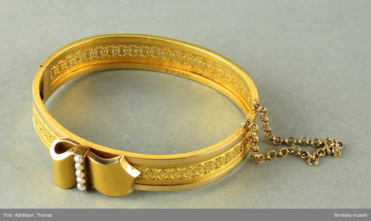 A) Armring i något oval form, 18 k guld. Dekoration, längsmed armringen, av  genombruten filigrandekor samt litet bandornament i guld med  5 st små, gråvita, satta äkta pärlor.
Armbandet är stelt och öpnas/slutes medelst "klick"-lås.
Säkerhetskedja vid låset.
Tillverkad 1888 av  Gustaf Dahlgren & Compani, Malmö.
Försedd med kattfot.

B) Etuie för armband. Hölje i mörkbrunfärgad kartong med tunn gulddekor utmed lockets övre kanter. Etuiet på insidan klätt med ljusblå sammet och siden. En löstagbar bottenplatta klädd i ljusblå sammet med en upphöjning för fästandet av armbandet (också den klädd i ljusblå sammet).

/Cecilia Wallquist 2019-03-29