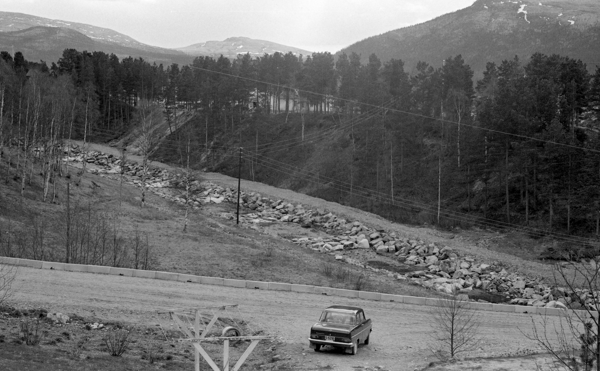 Fra Sandegga, som ligger «vestate» - altså vest for Glomma og kommunesenterert i Alvdal i Hedmark. Fotografiet er tatt fra et høyt punkt, muligens stillaset på Sandeggbakken, mot to veger. Den som går i forgrunnen, og som har kantstein av betong, later til å være etablert og velbrukt. Ut fra denne, fra høyre mot venstre i bildet, var man i ferd med å bygge en ny veg med grov stein i botnen og finere masser oppå. Vegbygginga må ha vært utført ved hjeøp av kraftige anleggsmaskiner. I forgrunnen, ved et tørketstativ uten klesvask, sto det en Opel kadett, antakelig en 1963-modell.