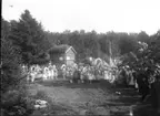 Folksamling, barn och vuxna vid Friluftsmuseet på Grännaberget, det är Hembygdsfest. Barnen håller blomsterbågar i händerna. De står framför pappersmakarbostället vars loftbod har en skylt: 