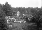 Folksamling, barn och vuxna vid Friluftsmuseet på Grännaberget, det är Hembygdsfest. Barnen håller blomsterbågar i händerna. De står framför pappersmakarbostället vars loftbod har en skylt: 