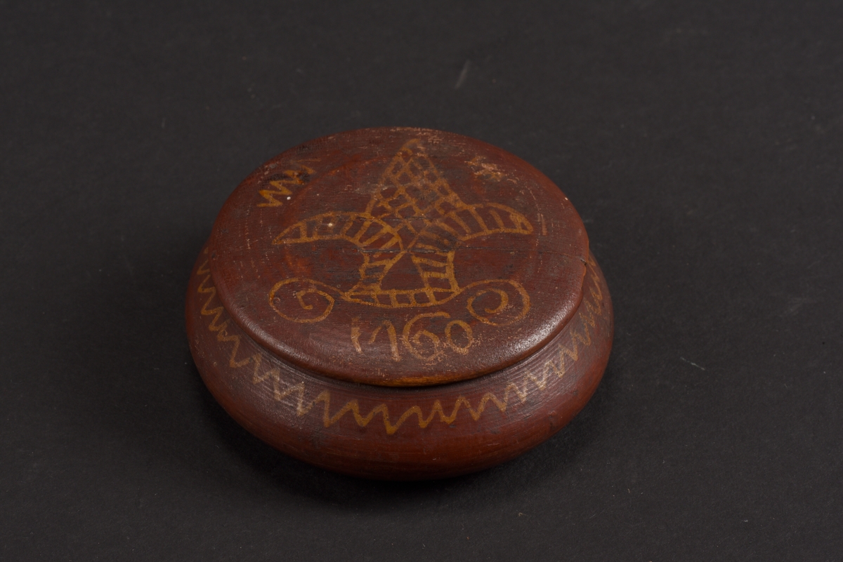 Rund, svarvad, ask med lock tillverkad av bok. Asken är målad på utsidan i rött med gula mönster; runt asken ett sicksackmönster och på locket ett årtal, 1760, samt en stiliserad lilja. Asken är skadad och en bit saknas.