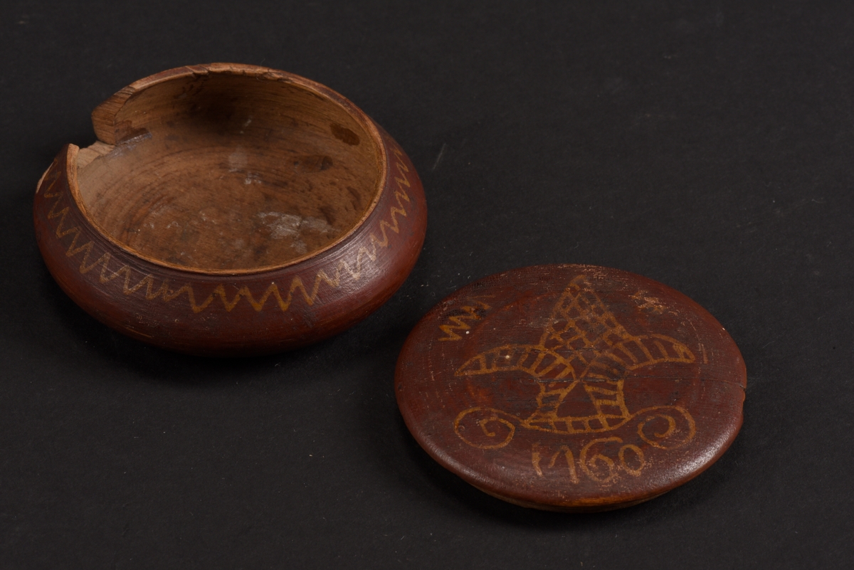 Rund, svarvad, ask med lock tillverkad av bok. Asken är målad på utsidan i rött med gula mönster; runt asken ett sicksackmönster och på locket ett årtal, 1760, samt en stiliserad lilja. Asken är skadad och en bit saknas.